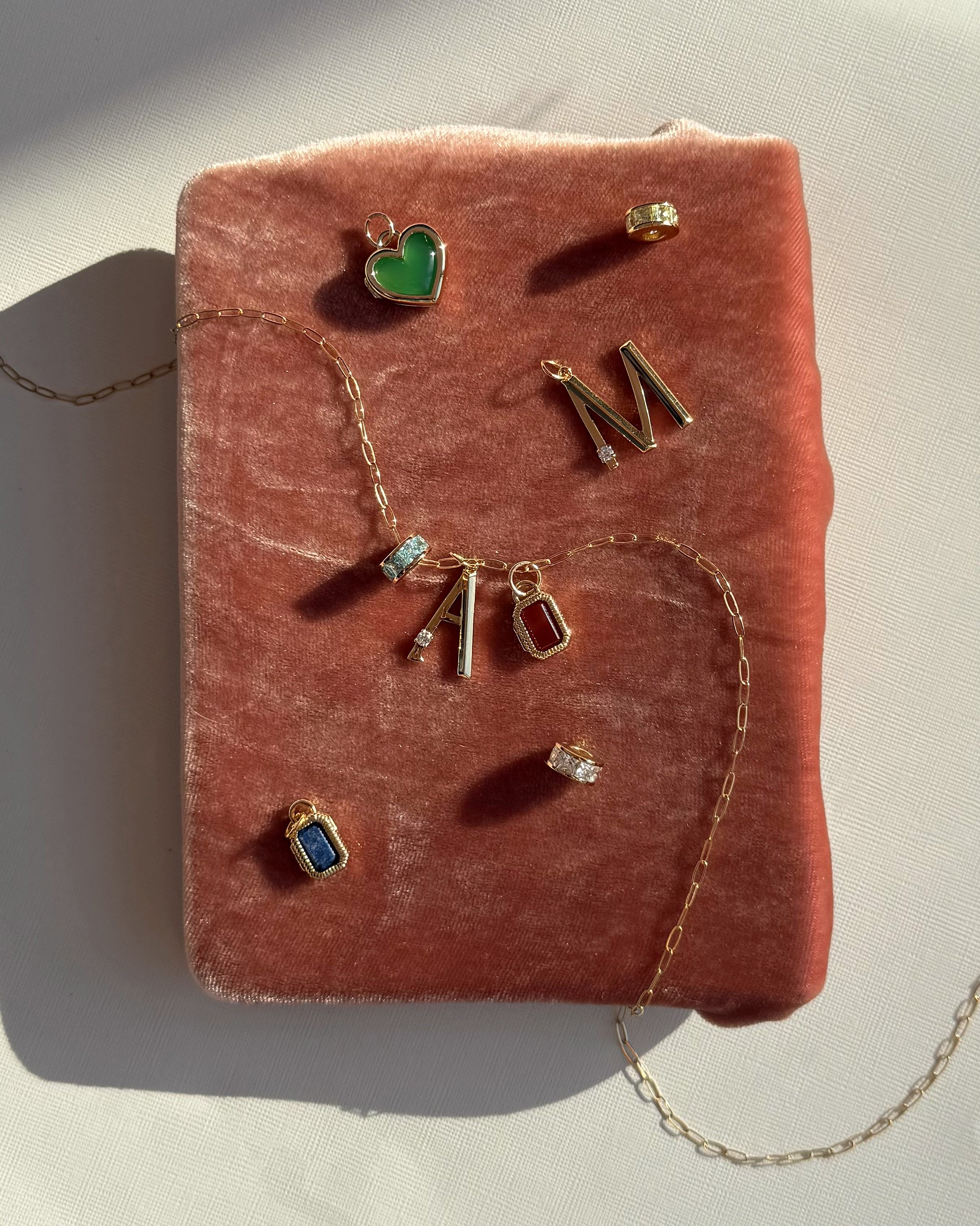Mod + Jo’s Este Birthstone Charm Spacer, gold plated necklace, personalized letter pendants A and M, and colored gemstone jewelry are displayed on a soft pink velvet surface in sunlight.