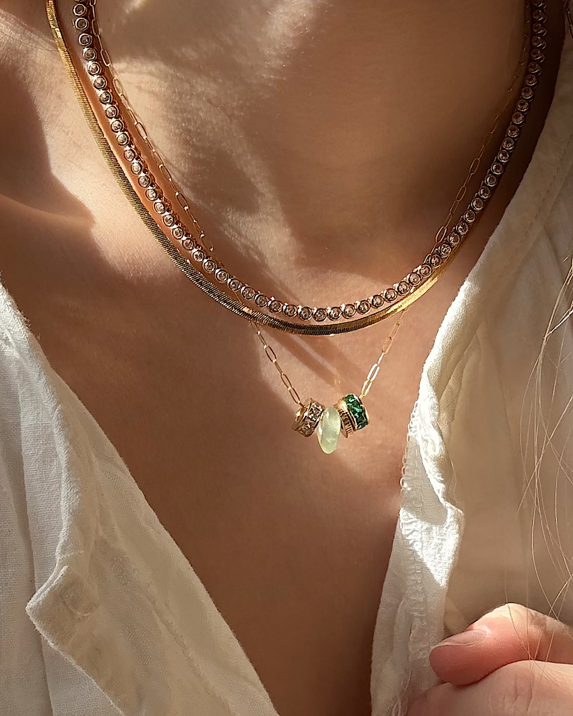 A close-up of a person wearing Mod + Jo’s Este Birthstone Charm Spacer—a gold necklace with three gemstone pendants—layered with a thick rhinestone choker over a textured white blouse, all highlighted by warm sunlight.