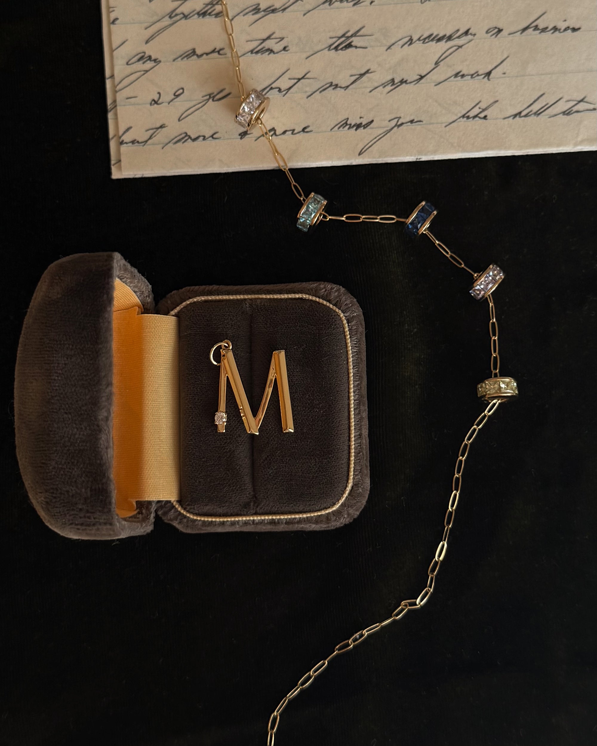 A Mod + Jo Este Birthstone Charm Spacer gold necklace with an “M” pendant is displayed in a velvet jewelry box on a black surface beside a handwritten letter.