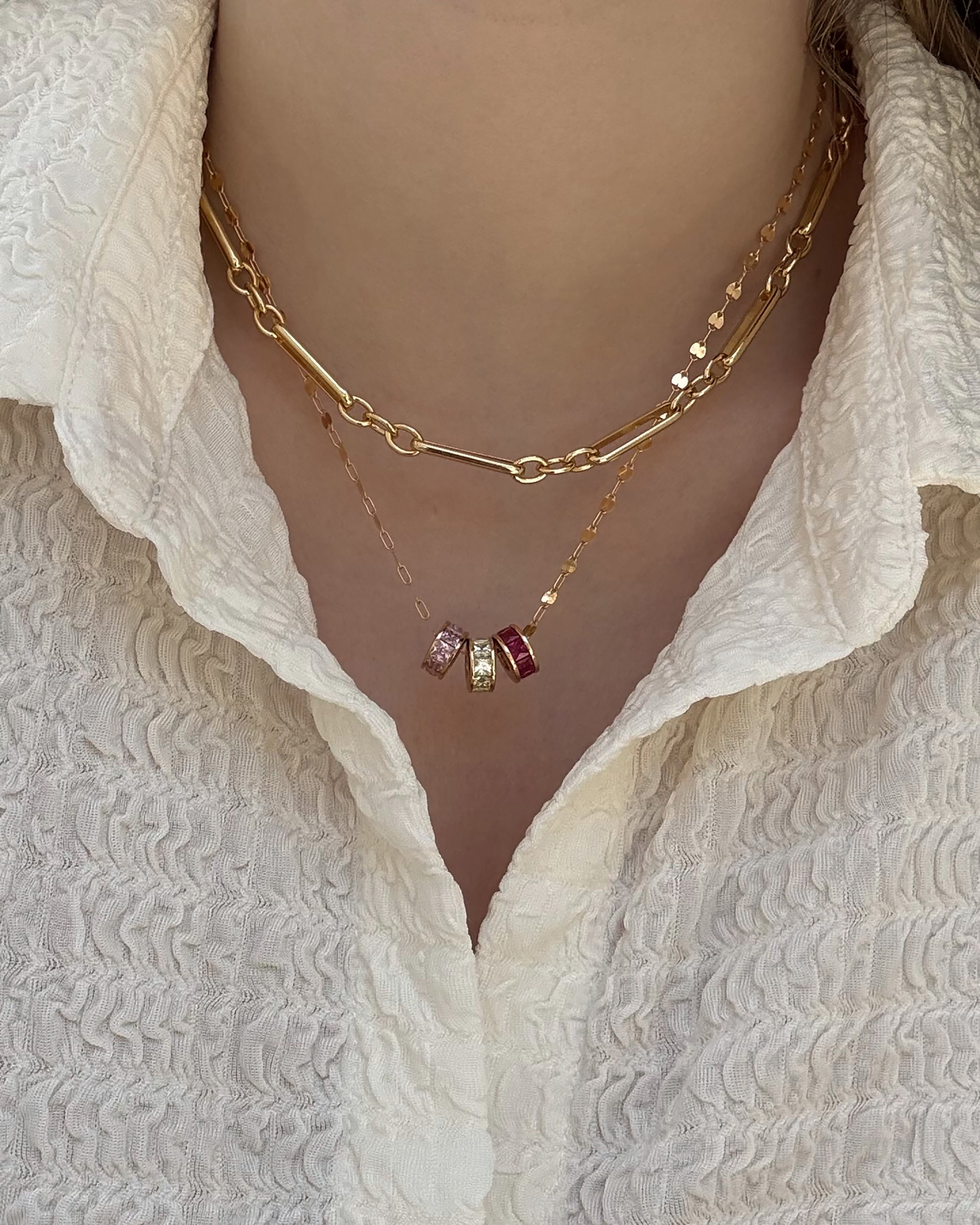 A person wears a textured white blouse with two Mod + Jo gold plated necklaces: a chunky chain and a fine chain featuring the Este Birthstone Charm Spacer with pink, clear, and red gemstone beads.