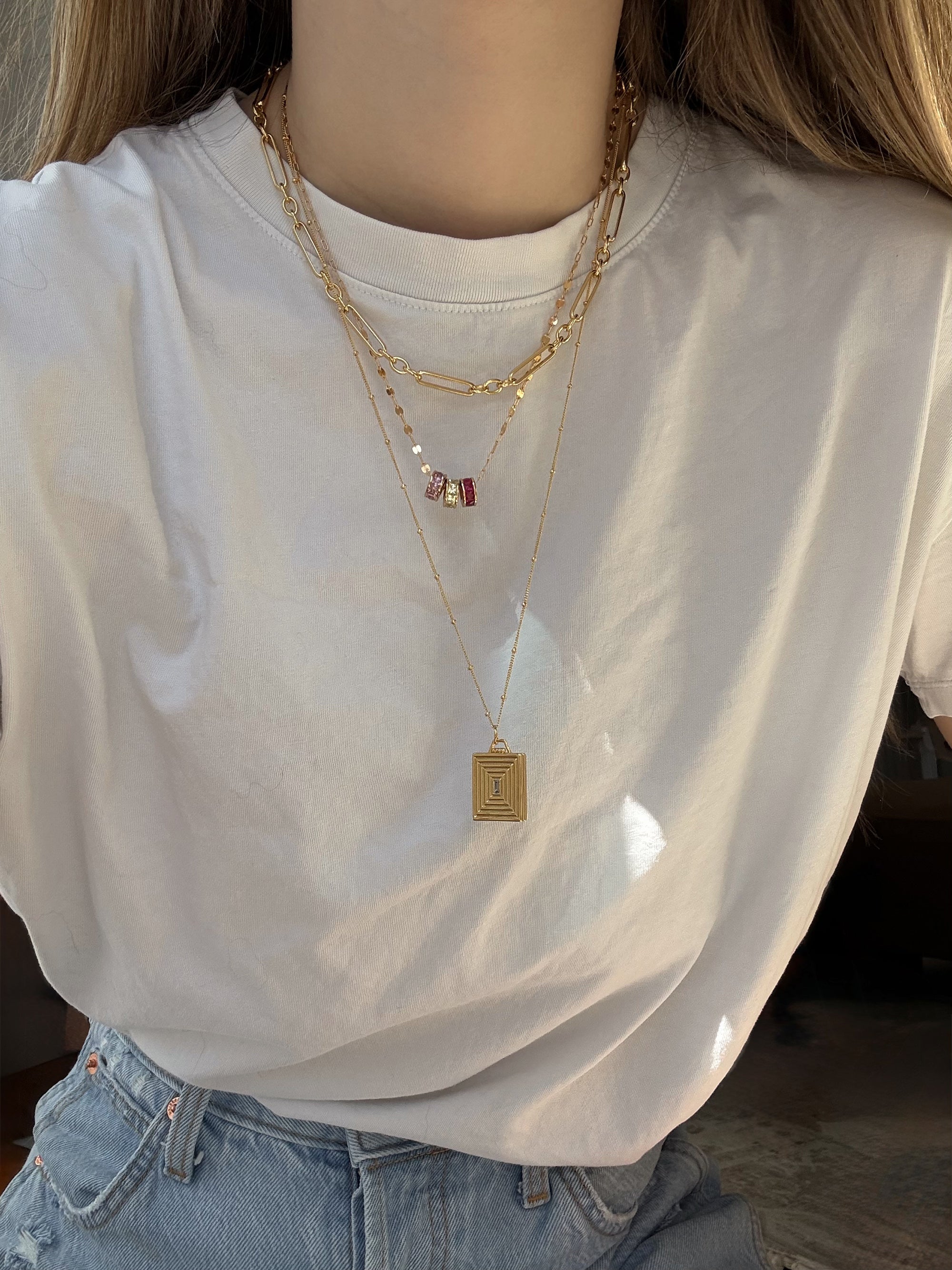 A person wearing a plain white t-shirt and light blue jeans, layered with three gold necklaces—one featuring the Mod + Jo Este Birthstone Charm Spacer for a personalized touch, plus two others: one with a rectangular pendant and one with small beads.