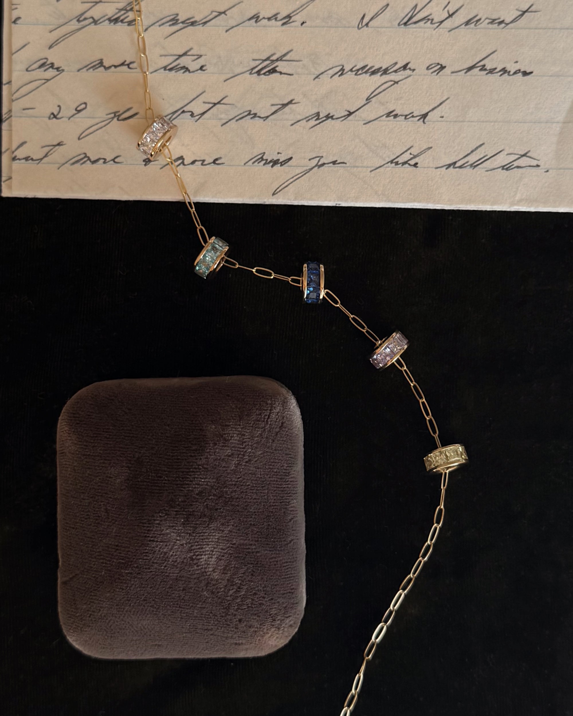 The Mod + Jo Este Birthstone Charm Spacer accents a gold chain with five jeweled rings, shown beside a closed black velvet jewelry box and a handwritten letter on lined paper.