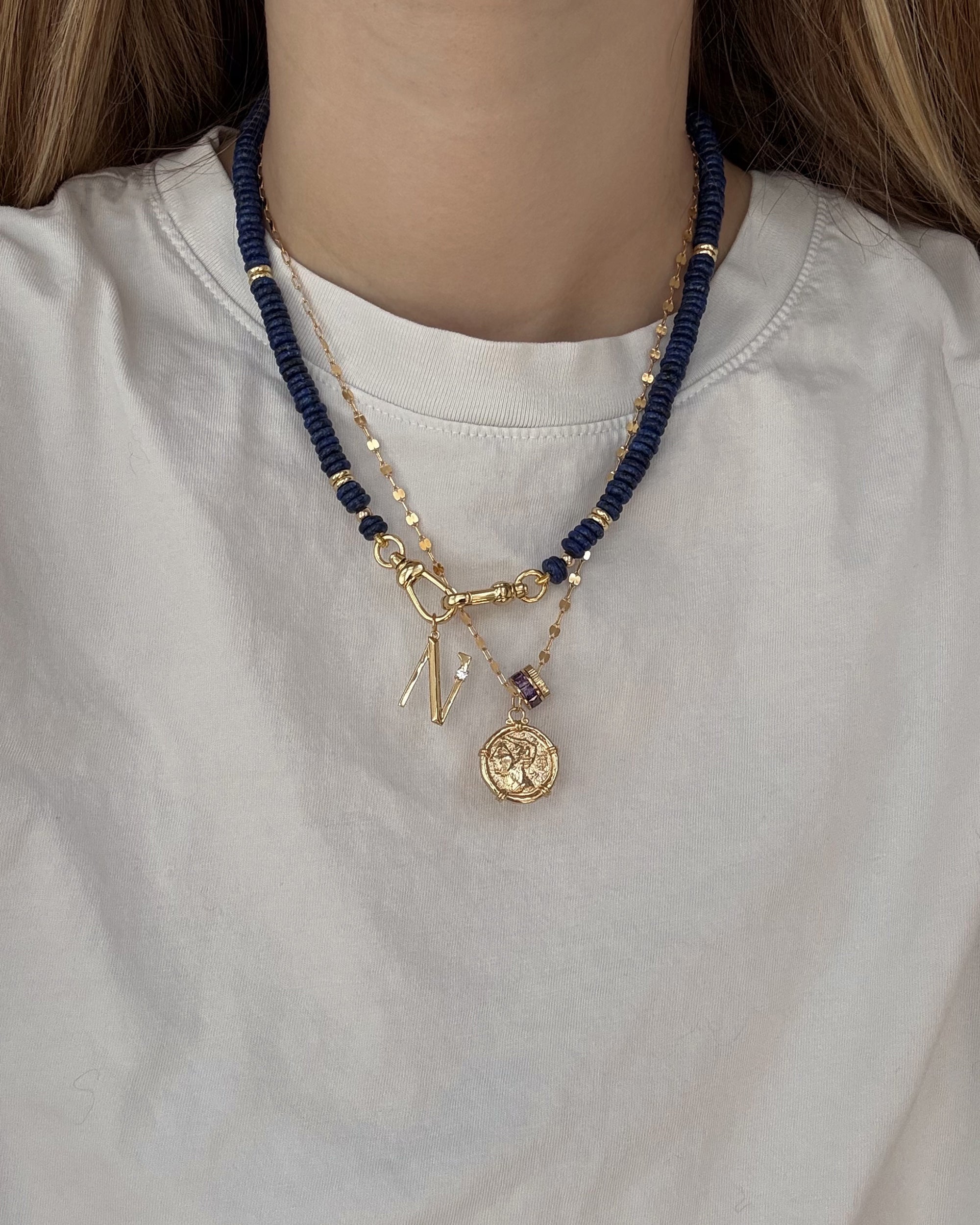 A person wears a white shirt with two layered necklaces: Mod + Jo's Eloise Letter Charm—a personalized gold initial and medallion—and a second necklace with dark blue beads and gold accents.