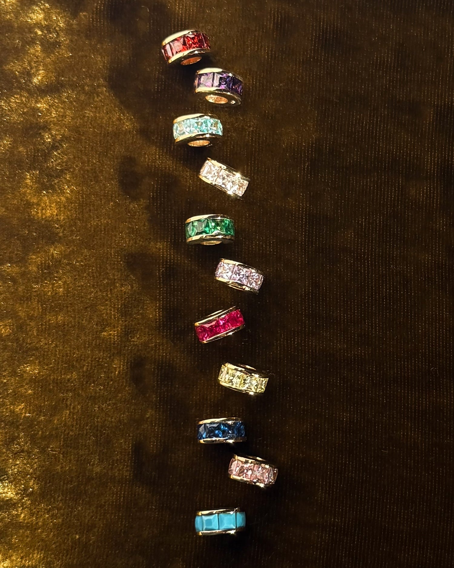 Ten Este Birthstone Charm Spacers by Mod + Jo, featuring vibrant rectangular center stones with side stones, are arranged vertically on a dark brown surface—ideal for adding color and elegance to personalized necklaces.