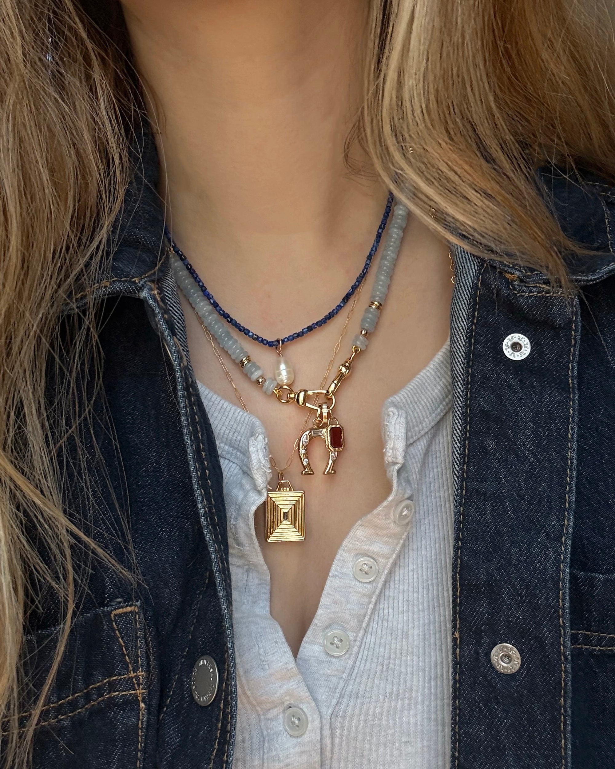 A person wears Mod + Jo’s Sonia Gemstone Necklace—featuring aquamarine beads—layered with gold jewelry and a gray beaded strand over a white top and dark denim jacket.