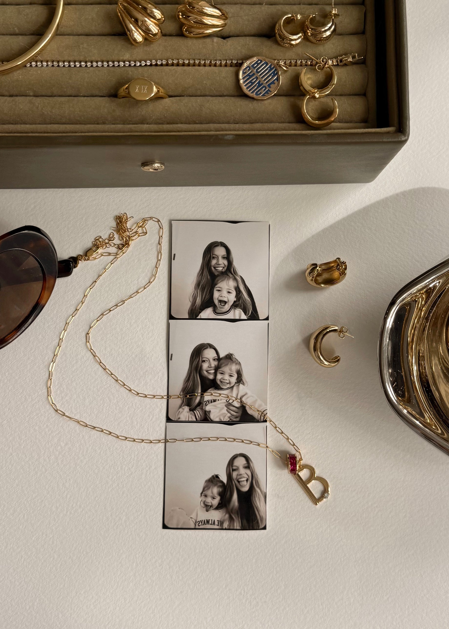 A red velvet jewelry box holds gold hoop earrings and a personalized Mod + Jo Eloise Letter Charm necklace with an S initial and star pendant. Another gold letter charm necklace and glass bottles rest on a white surface.
