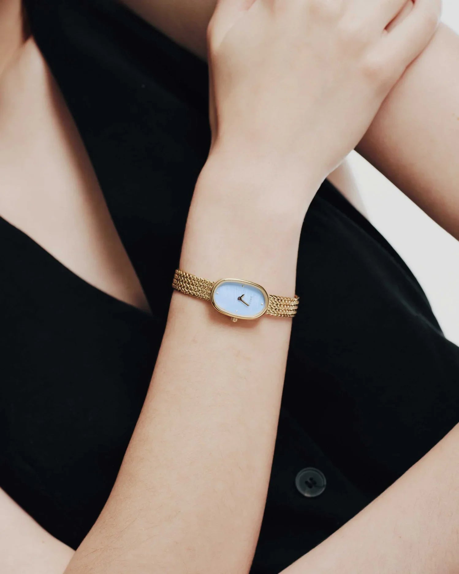 A person in a sleeveless black top showcases the Jane Watch Small (Tethered) by Breda, featuring a gold-plated band and rectangular light blue face, on their left wrist, while their right arm rests across their body.