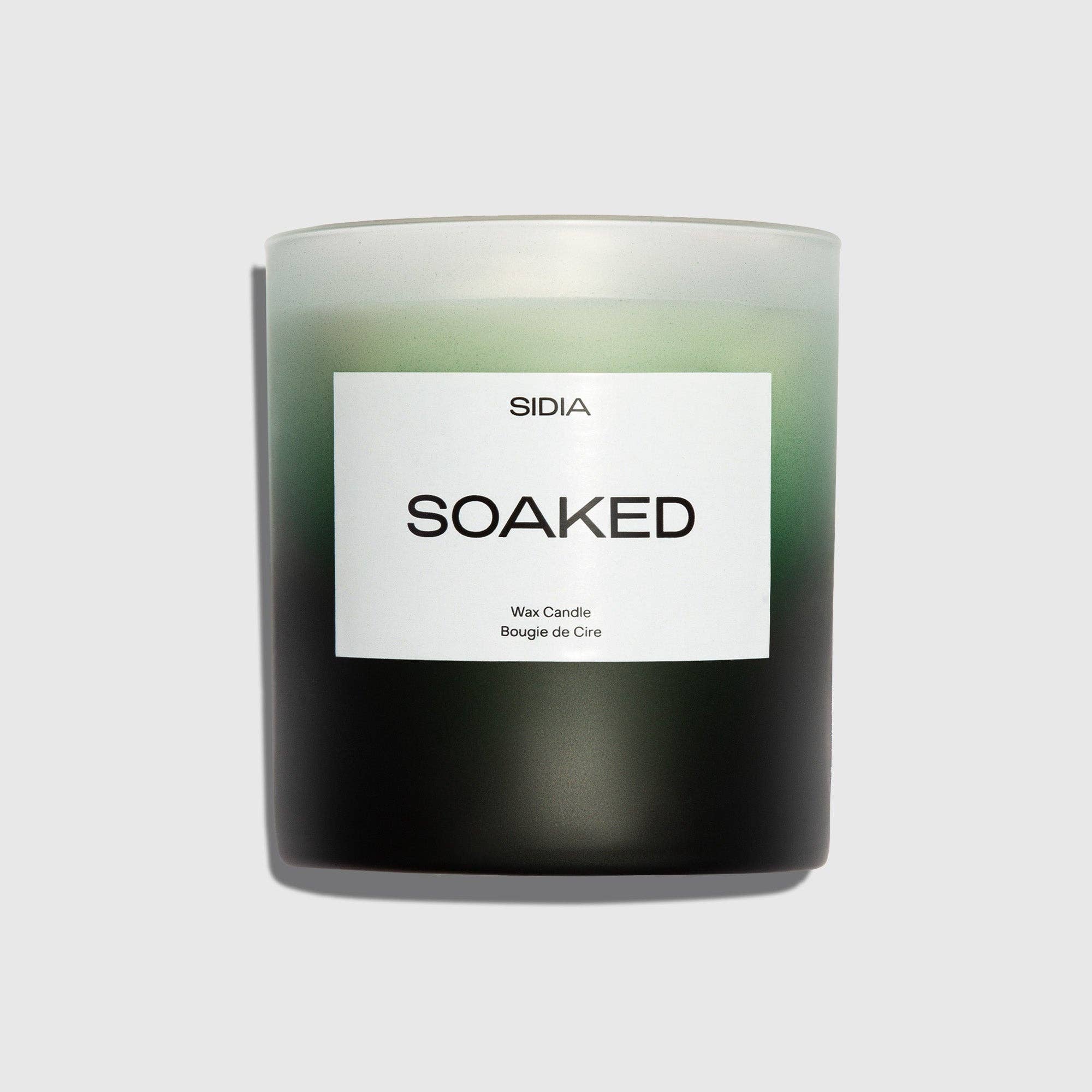 A black and green SOAKED Candle by SIDIA, housed in a frosted glass container with a white label, sits against a plain light gray background. Its subtle notes of juniper and jasmine evoke the refreshing scent of wild herbs.