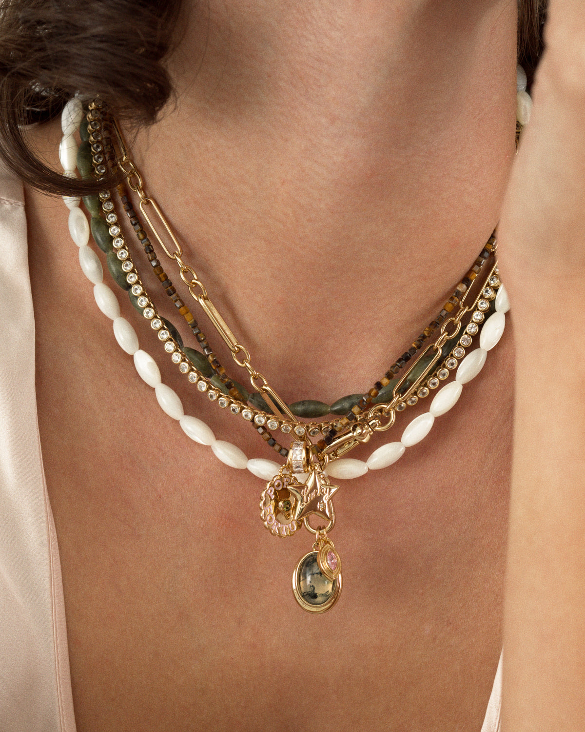 The Mod + Jo Sutton Gemstone Necklace is an 18″ layered set featuring two strands of white rice-shaped mother of pearl, a gold link chain, a strand of dark brown beads, a strand of clear rhinestones, and several small gold charms.