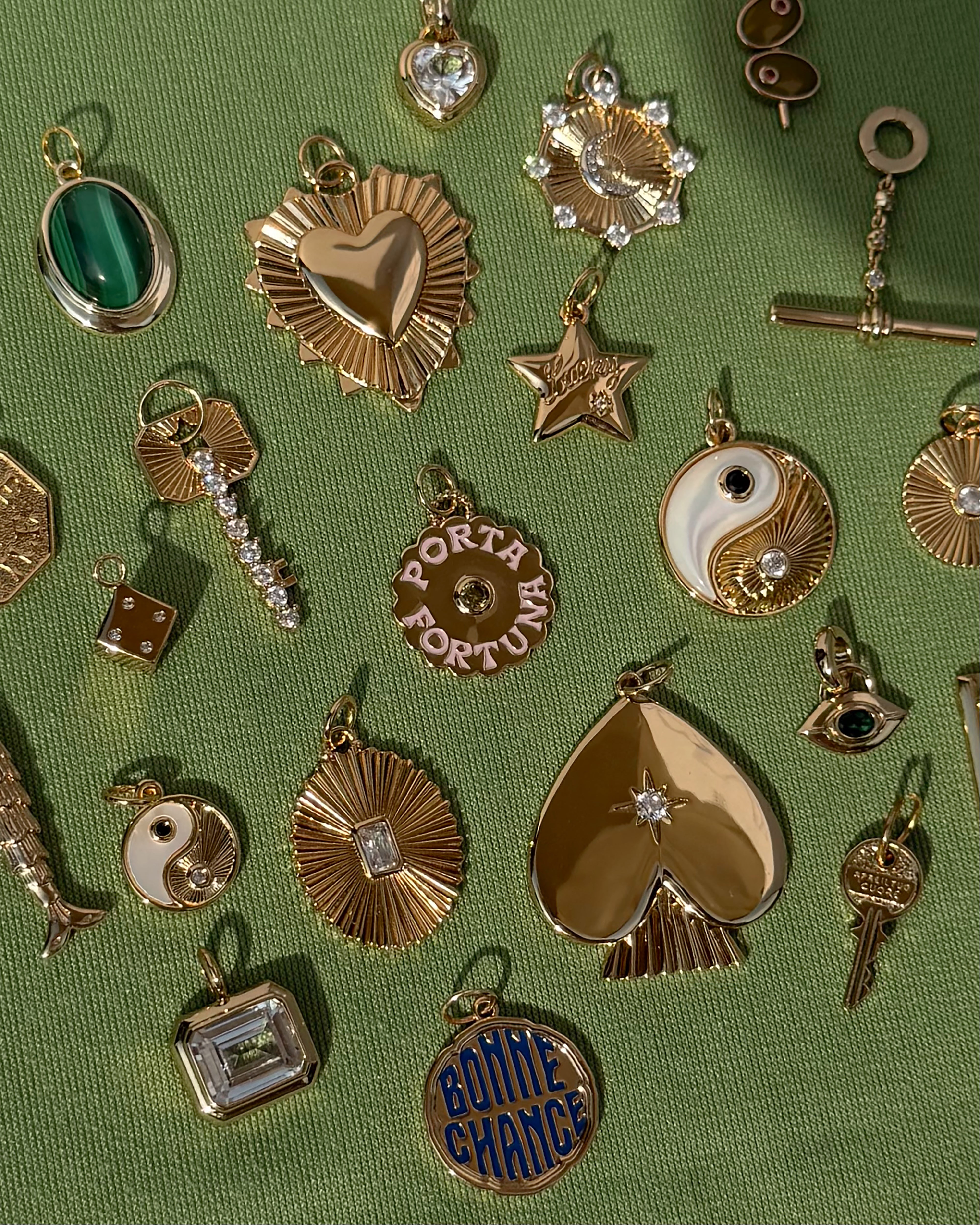 Mod + Jo gold and jeweled charms—including hearts, stars, yin-yangs, a protective pendant, and the Nyx Evil Eye Charm—are displayed on a green textured fabric background.