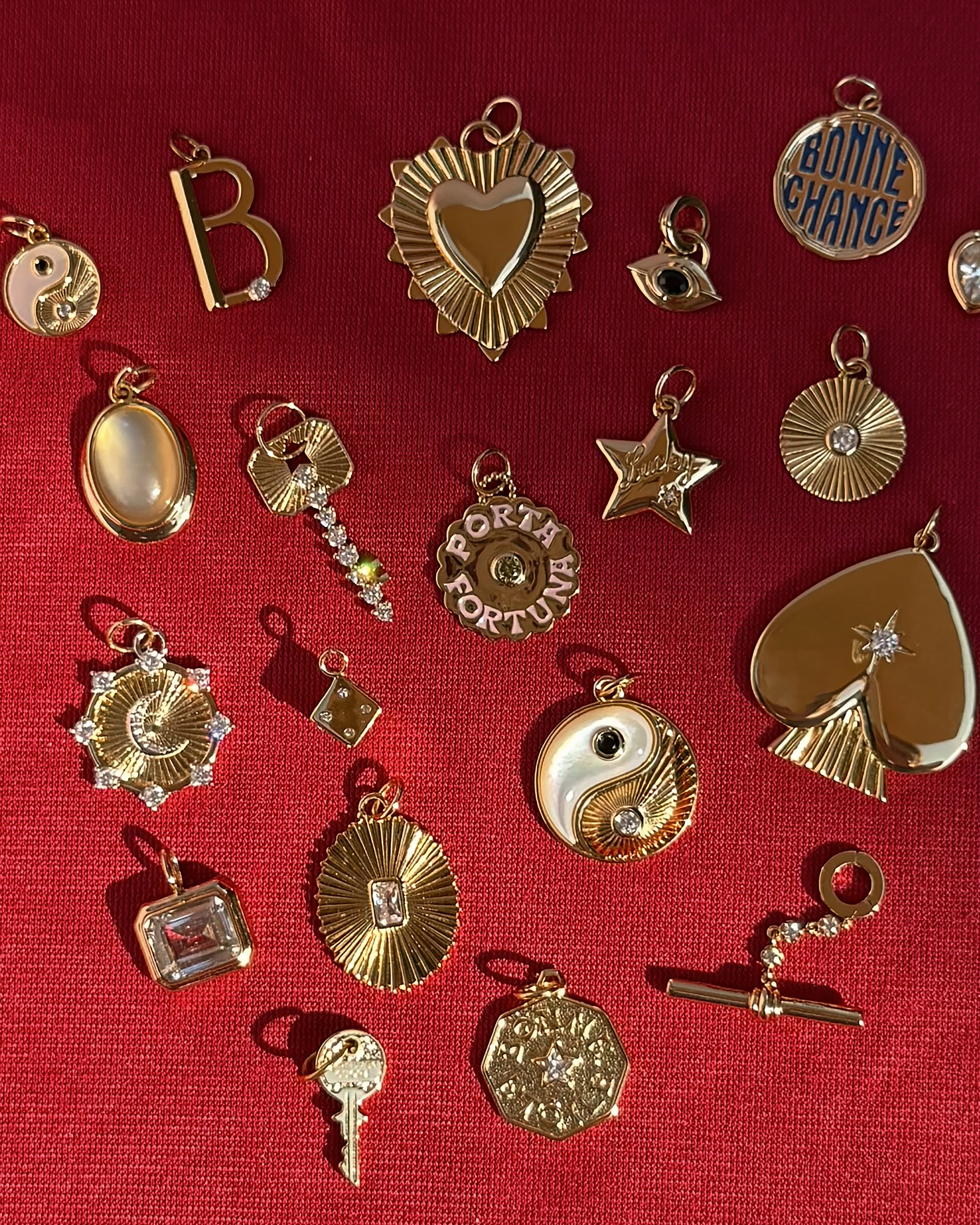 A variety of gold and jeweled charms—including stars, hearts, keys, a yin-yang, an oval reading BONNE CHANCE, and the shimmering Mod + Jo Bonne Etoile Charm reminiscent of vintage gold coin jewelry—are arranged on a textured red fabric background.