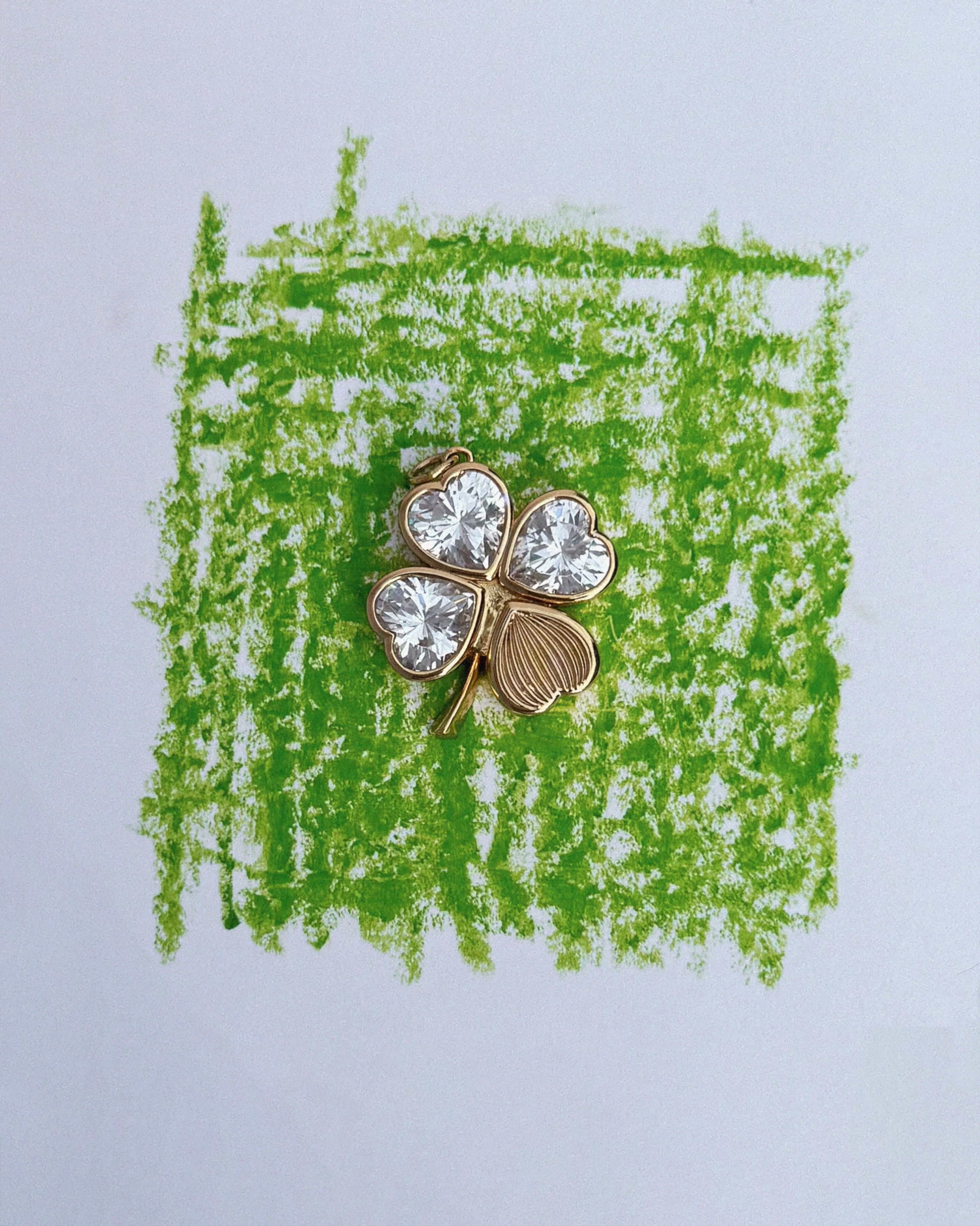 A gold Perry Four-Leaf Clover Locket Charm by Mod + Jo, featuring three leaves adorned with clear gemstones, rests on a green crayon square drawn on white paper, evoking the whimsy and elegance of cherished keepsakes.
