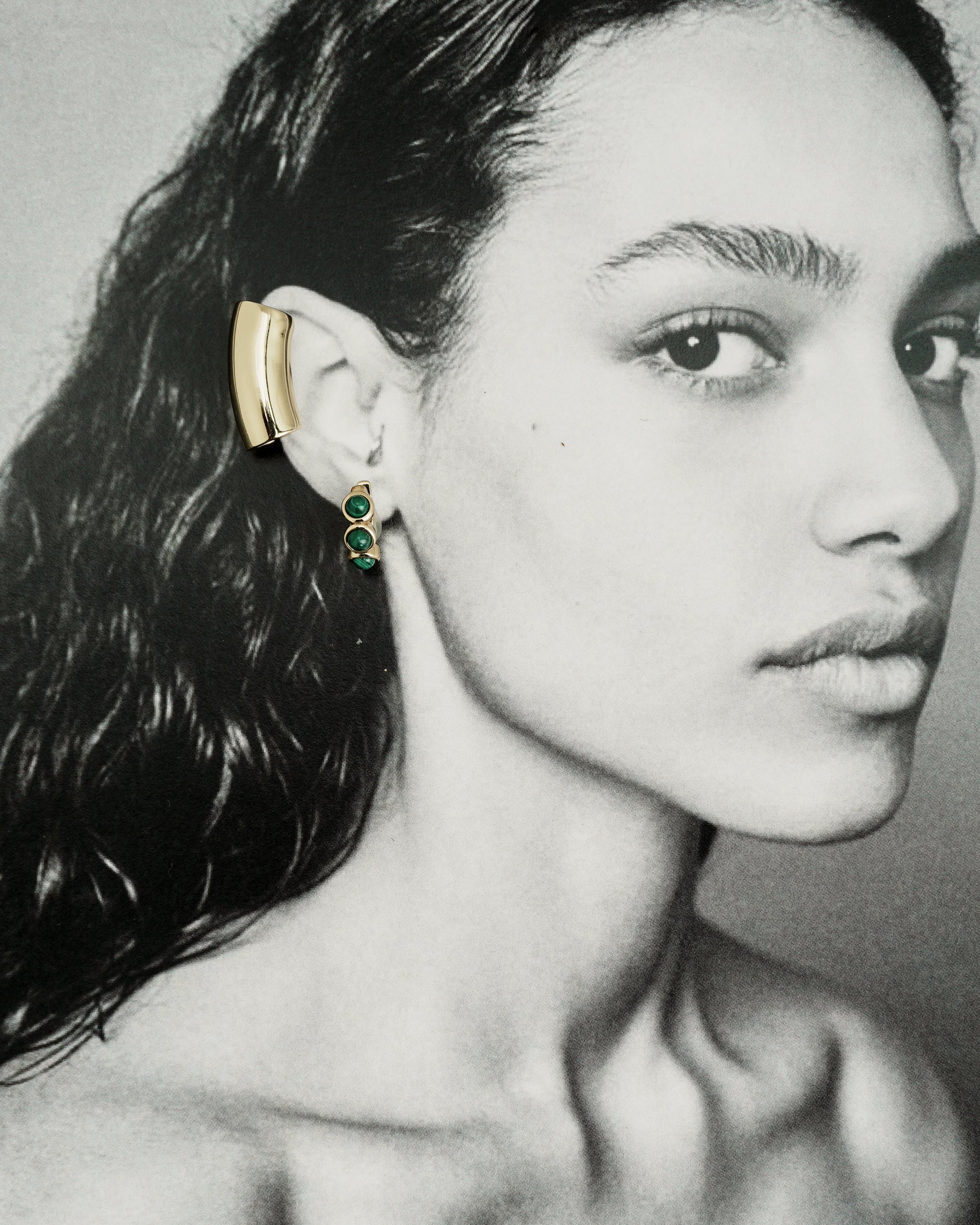 Black-and-white portrait of a woman with wet, wavy hair, wearing the *Preorder* Lennon Tube Helix Ear Cuff by Mod + Jo—a large, minimalist 18k gold piece—alongside a dangling green stone earring. She gazes calmly at the camera.