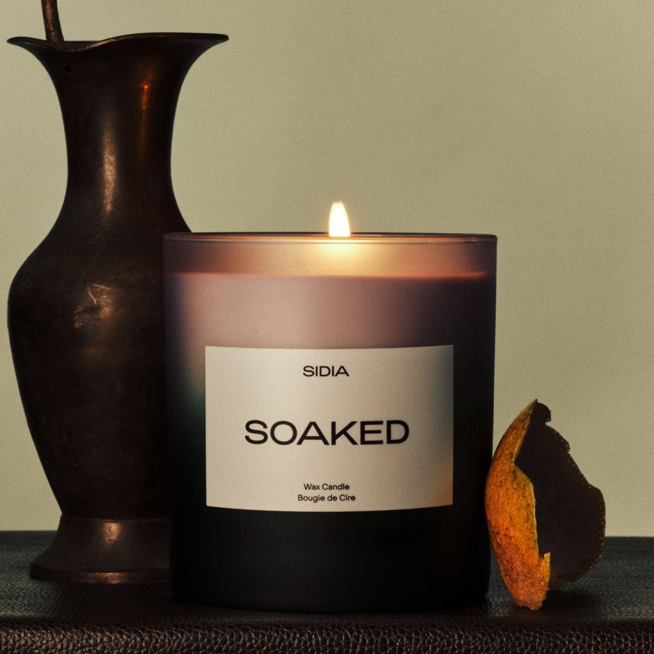 A glowing SOAKED Candle by SIDIA rests next to a dark vase and a piece of dried orange peel, arranged on a textured surface against a plain backdrop, releasing delicate hints of jasmine and herb into the air.