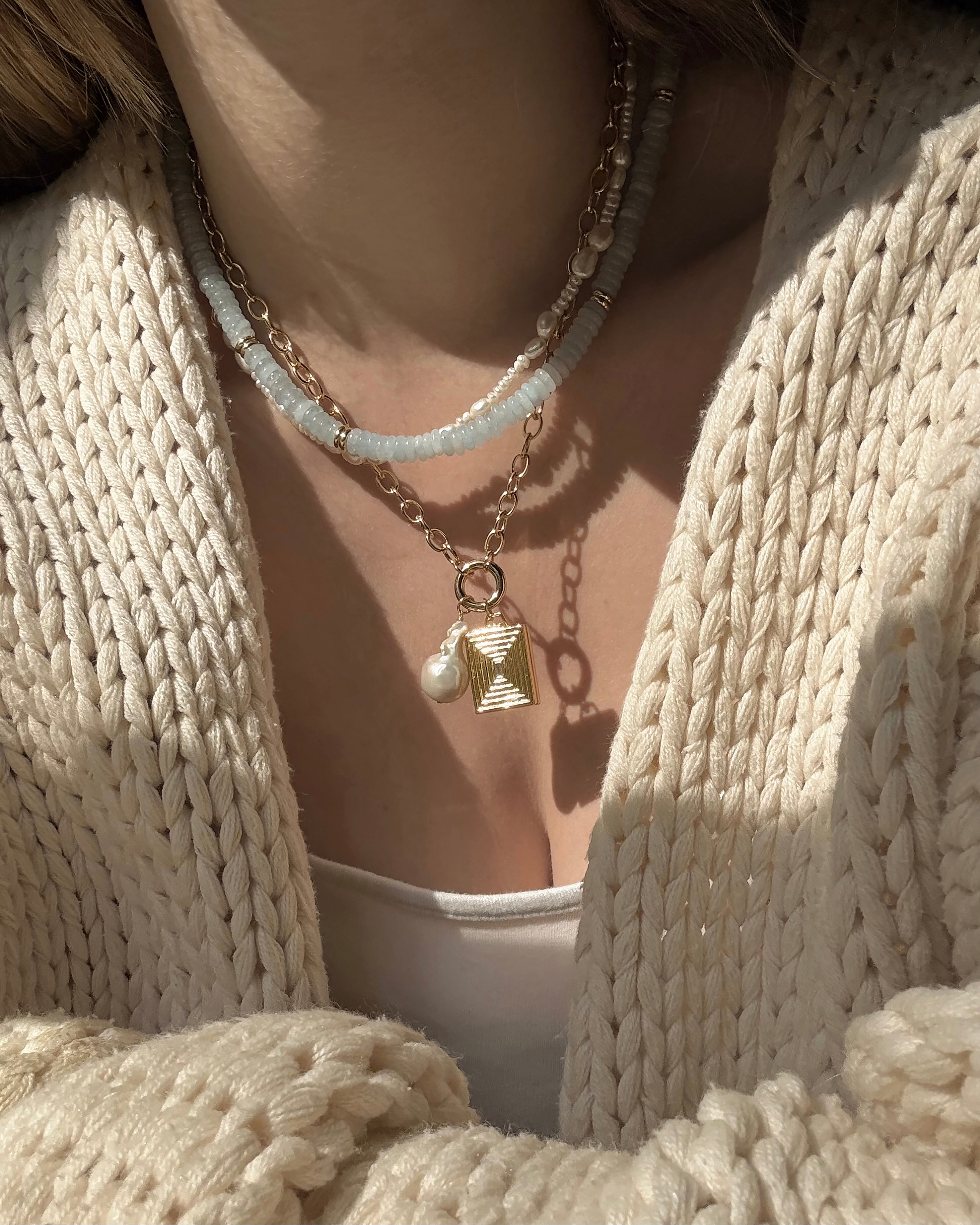 A person wears a chunky cream knit cardigan and white top, layered with three necklaces: Mod + Jo’s Presley Charm Clip Necklace, a gold chain with textured pendant and pearl, and a blue beaded necklace.