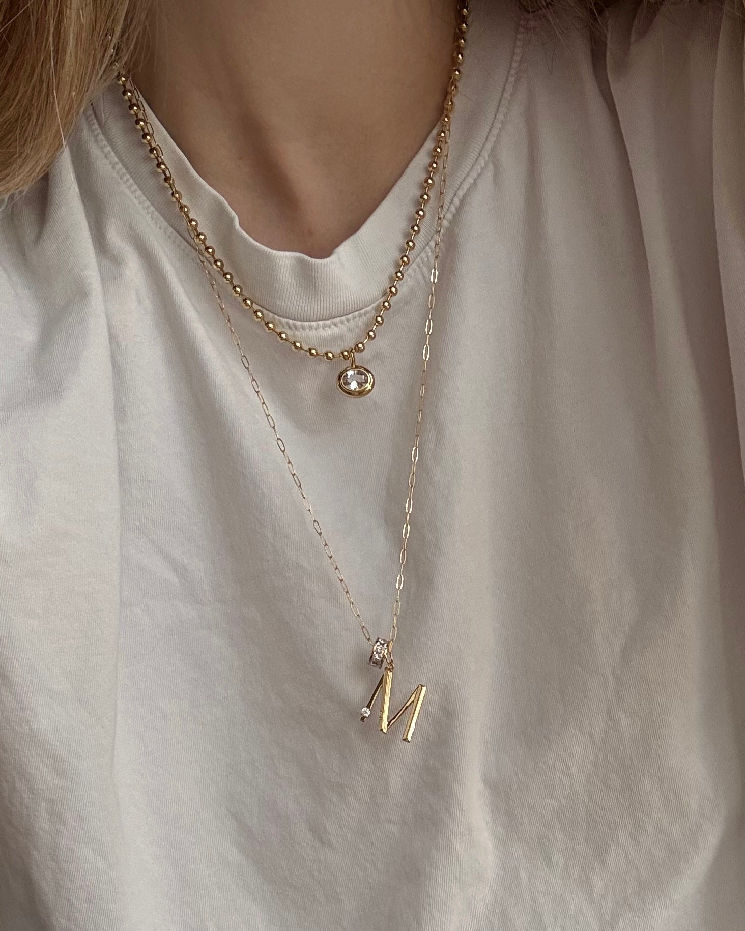 A person wears a plain white t-shirt and two gold necklaces: one is the Mod + Jo Wylder Paperclip Chain Necklace with a small round pendant, and the other has a larger letter M pendant.