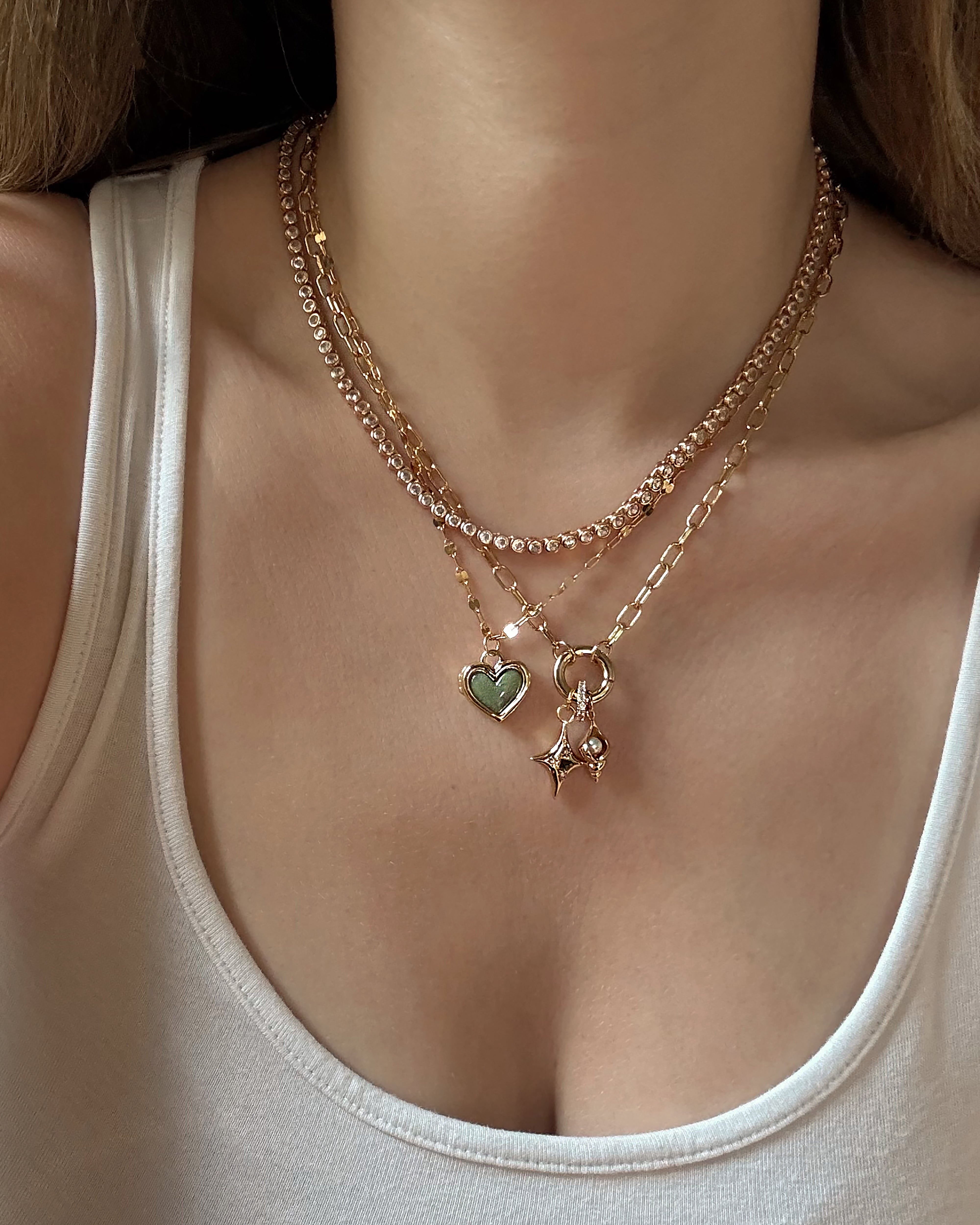 A person in a white tank top layers three gold necklaces, including the Mod + Jo Darcy Charm Clip Necklace, featuring 14k gold plating with a green heart pendant, a star-shaped charm, and one made of small gold links.