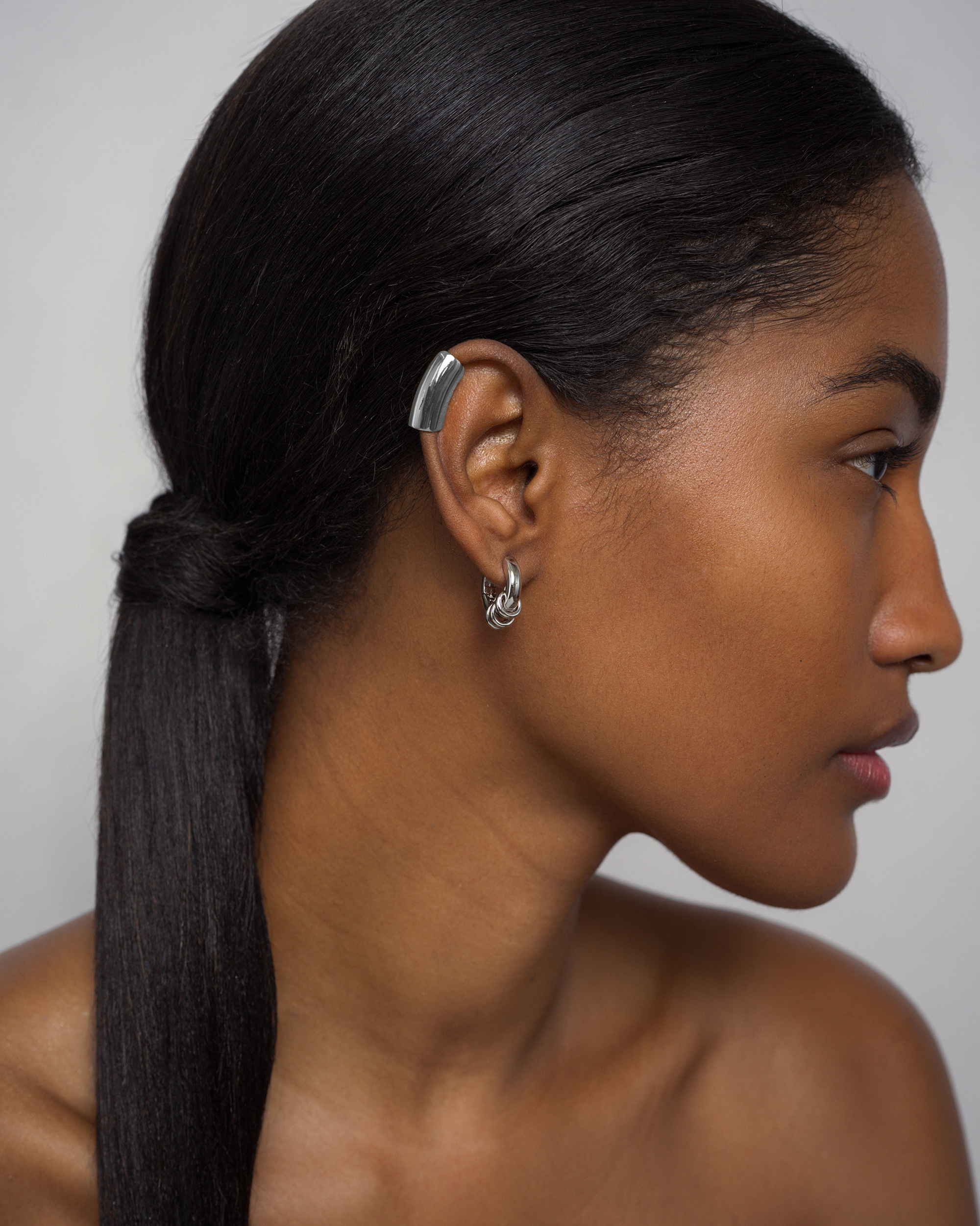 A close-up side profile of a woman with long, straight black hair wearing a silver curved ear cuff and a small silver hoop earring, against a light gray background.