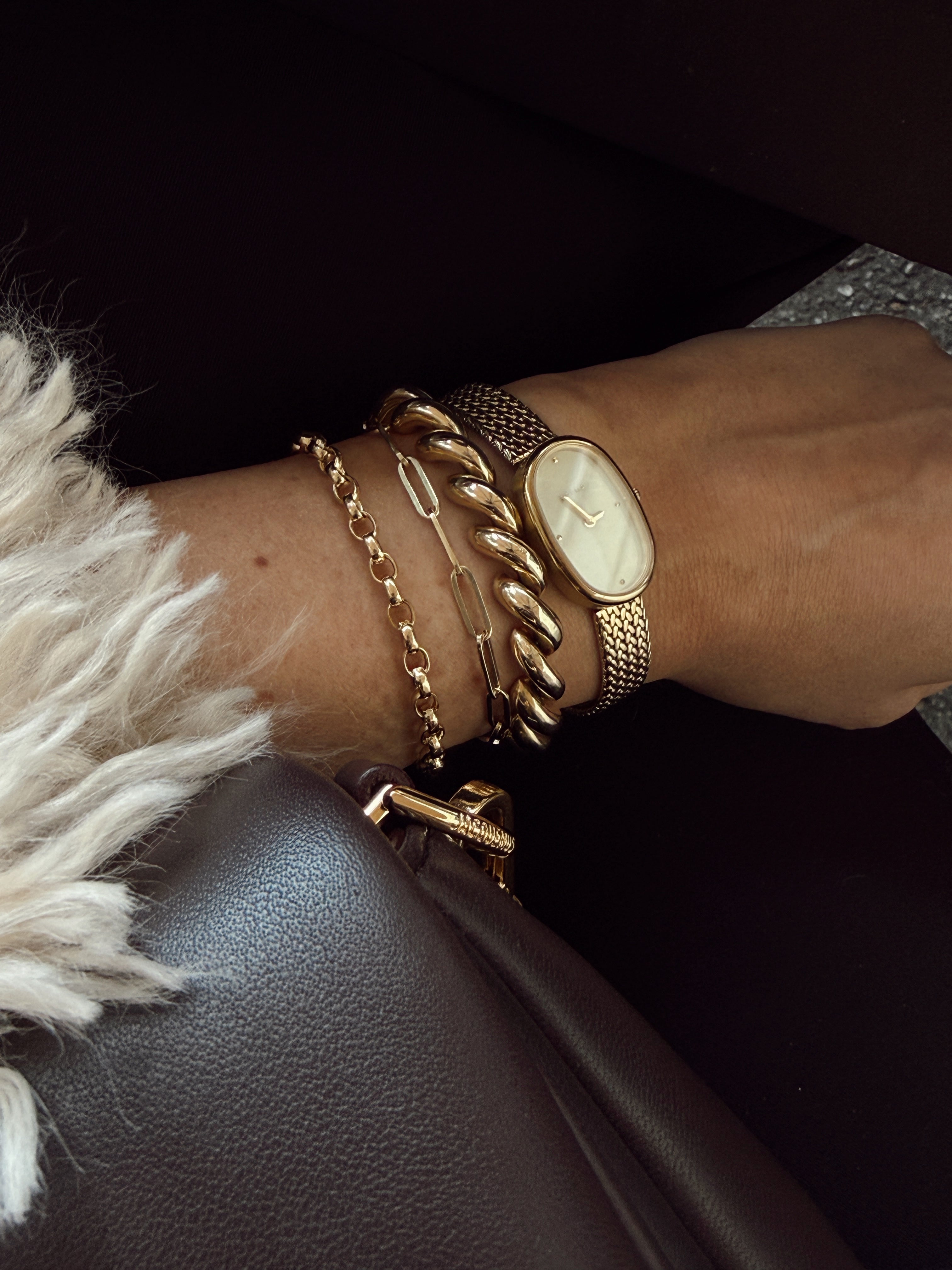 A close-up of a wrist adorned with three gold bracelets and a gold wristwatch with an oval face. The person is wearing dark clothing and a beige fur-trimmed sleeve. A brown leather bag is partially visible.
