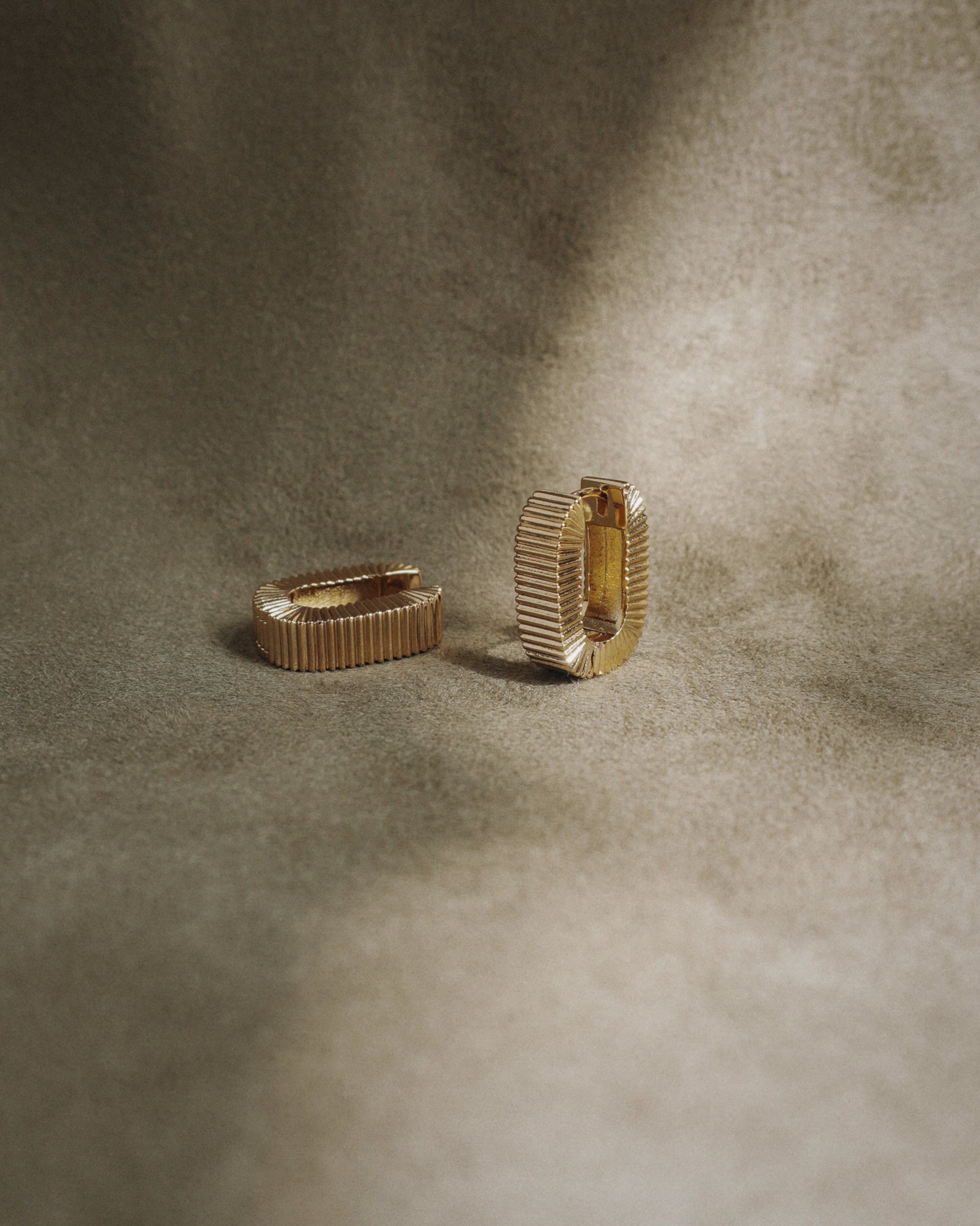 The Elly Fluted Hoops by Mod + Jo, featuring a gold textured design, are displayed on a soft brownish-beige fabric background with gentle lighting.
