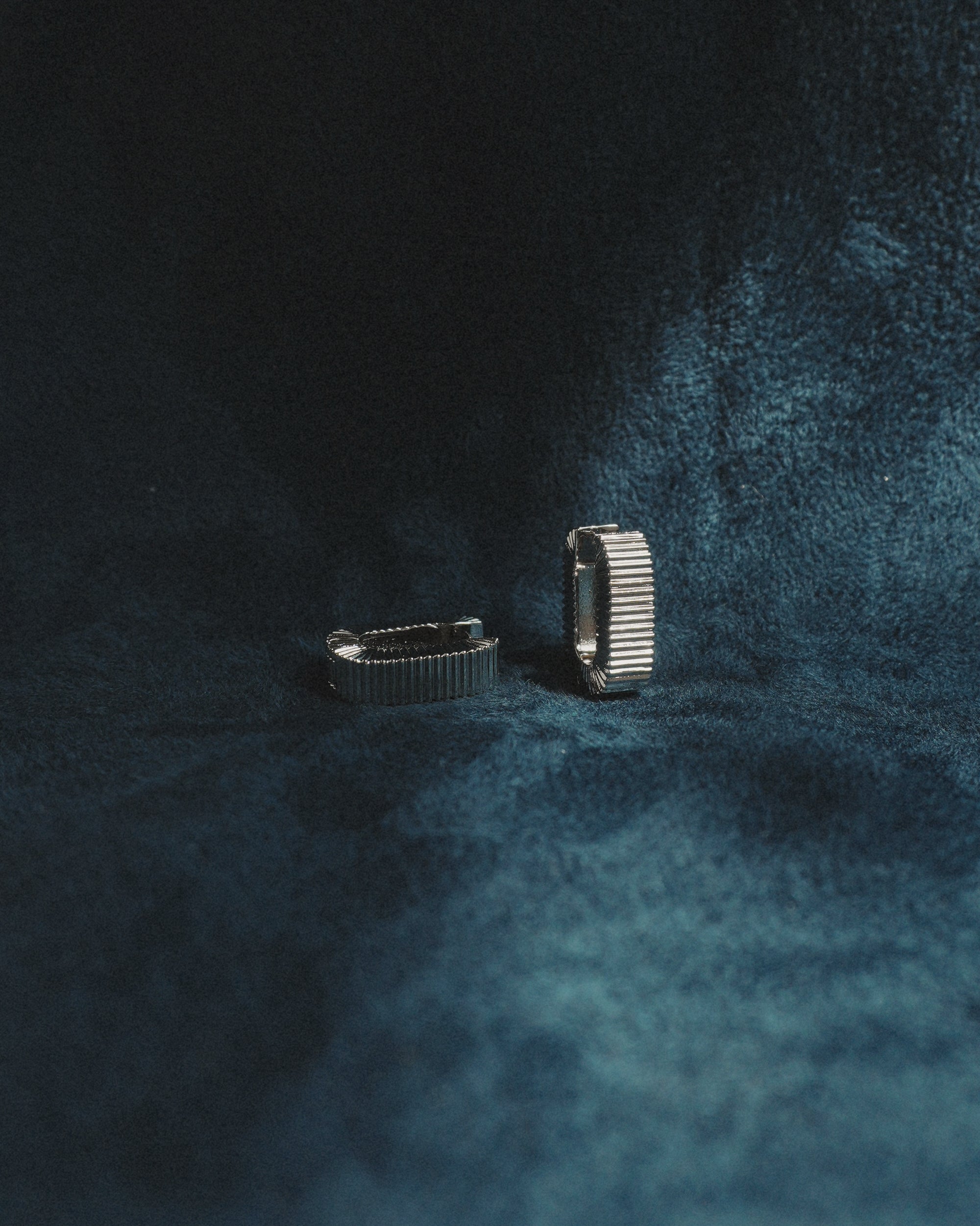 A pair of Elly Fluted Hoops by Mod + Jo, crafted in silver, are displayed upright on a dark blue, velvet-textured surface, with soft lighting casting gentle shadows around them.