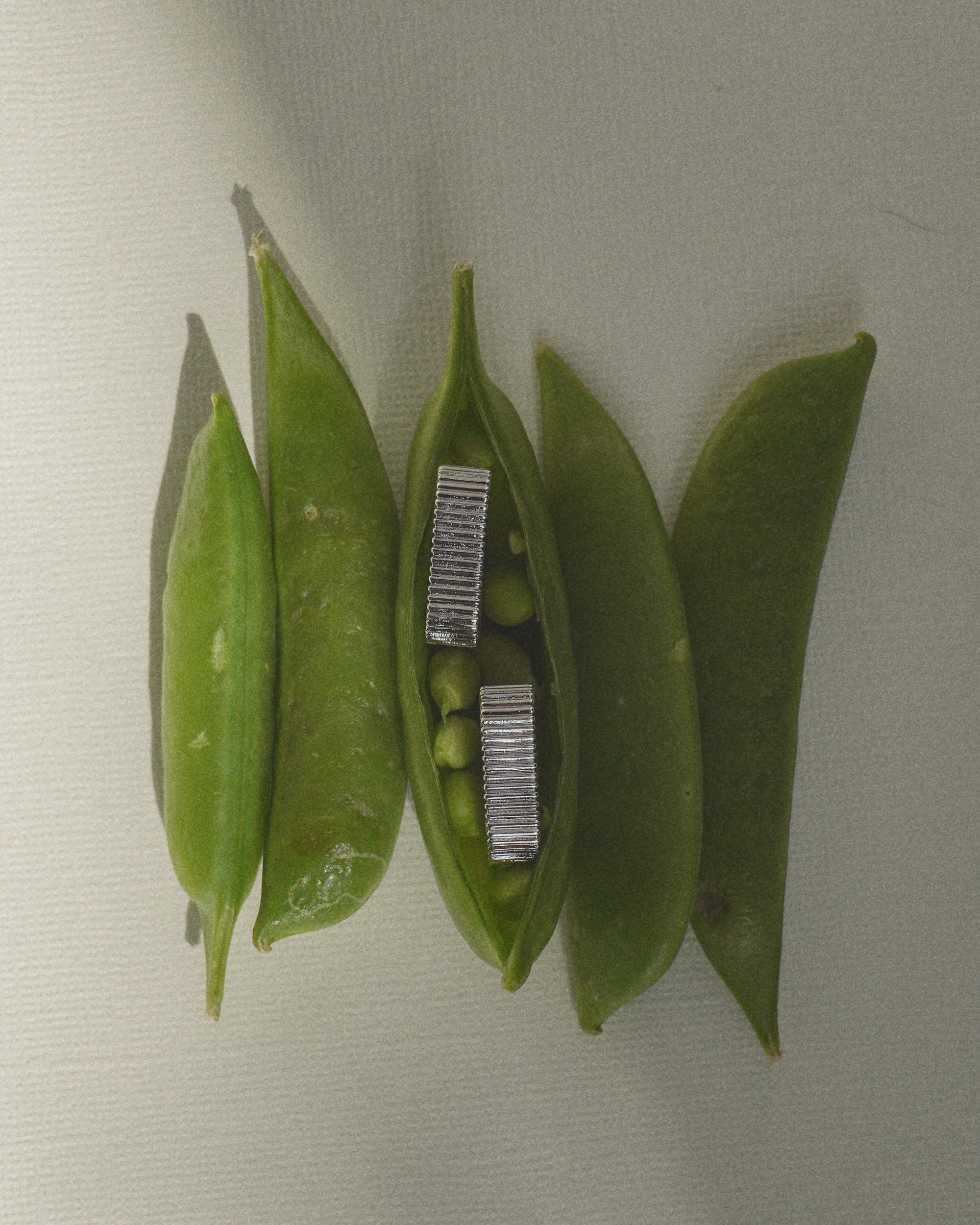 Four green pea pods rest on a light surface; one open pod reveals small peas with a pair of Mod + Jo’s Elly Fluted Hoops nestled inside.