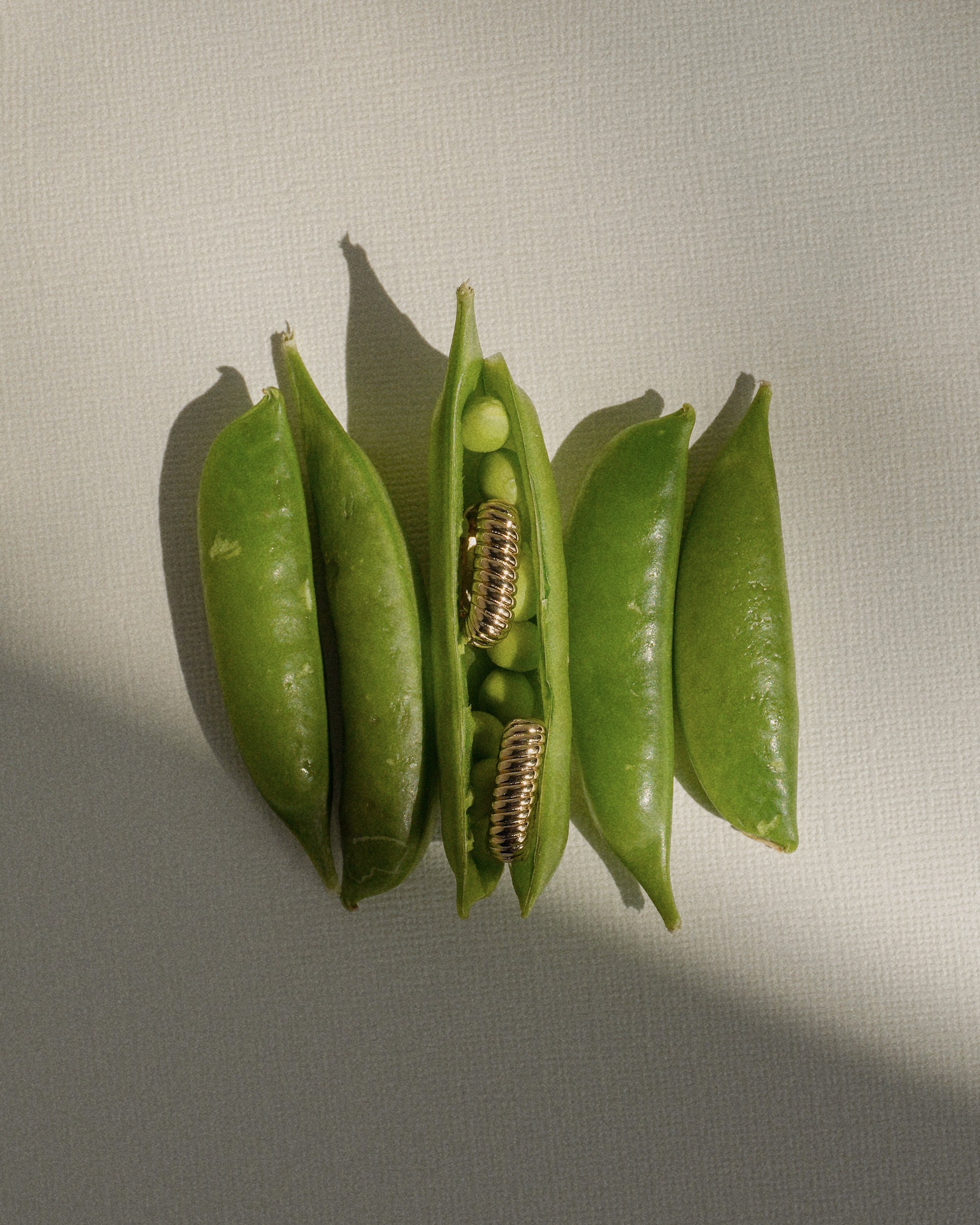Four pea pods rest on a textured surface; the middle pod is open, revealing two golden screw coils nestled among the peas—an artful fusion of nature and design reminiscent of Mod + Jo’s Elly French Rope Hoops.
