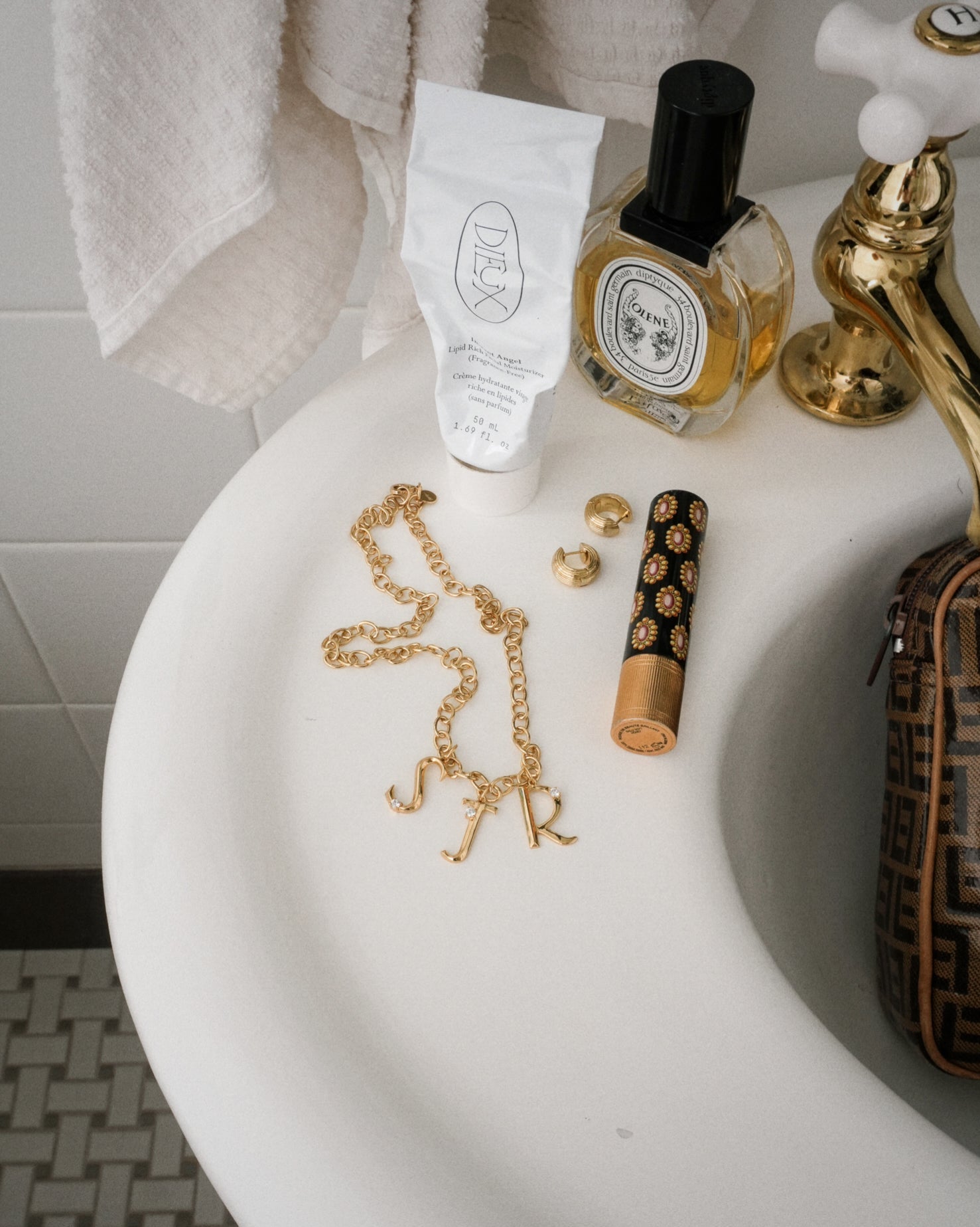 A Mod + Jo Eloise Letter Charm, gold jewelry, hand cream, perfume, decorative lipstick, and a small brown-patterned bag sit on the bathroom sink counter. A towel hangs above the sink.