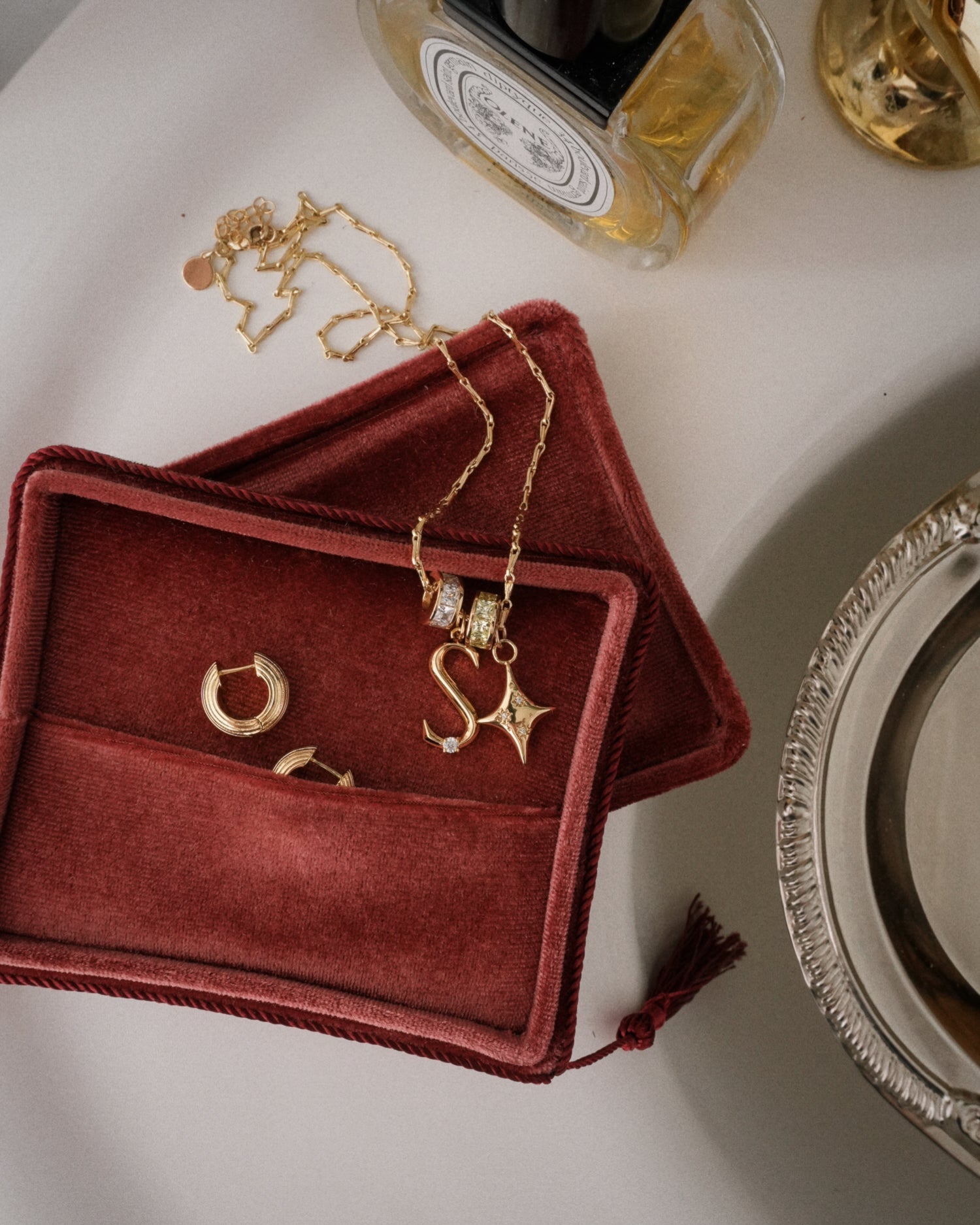 A red velvet jewelry box holds gold hoop earrings and a personalized Mod + Jo Eloise Letter Charm necklace with an S initial and star pendant. Another gold letter charm necklace and glass bottles rest on a white surface.