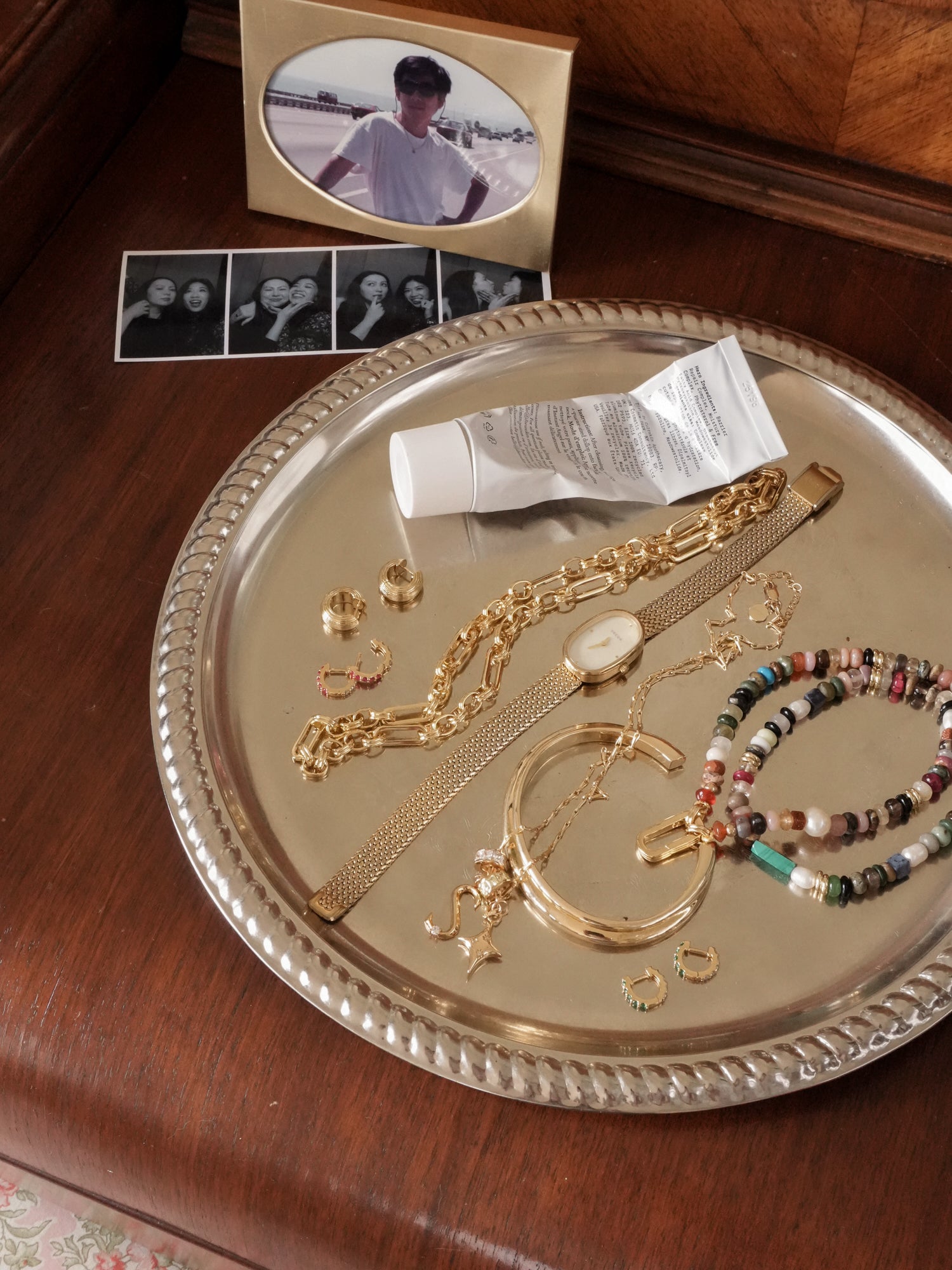 A silver tray on a wooden surface displays gold jewelry, the Mod + Jo Andy Gemstone Necklace, a beaded bracelet, carabiner clasp earrings, a wristwatch, and cream. Behind it are framed photos of people.