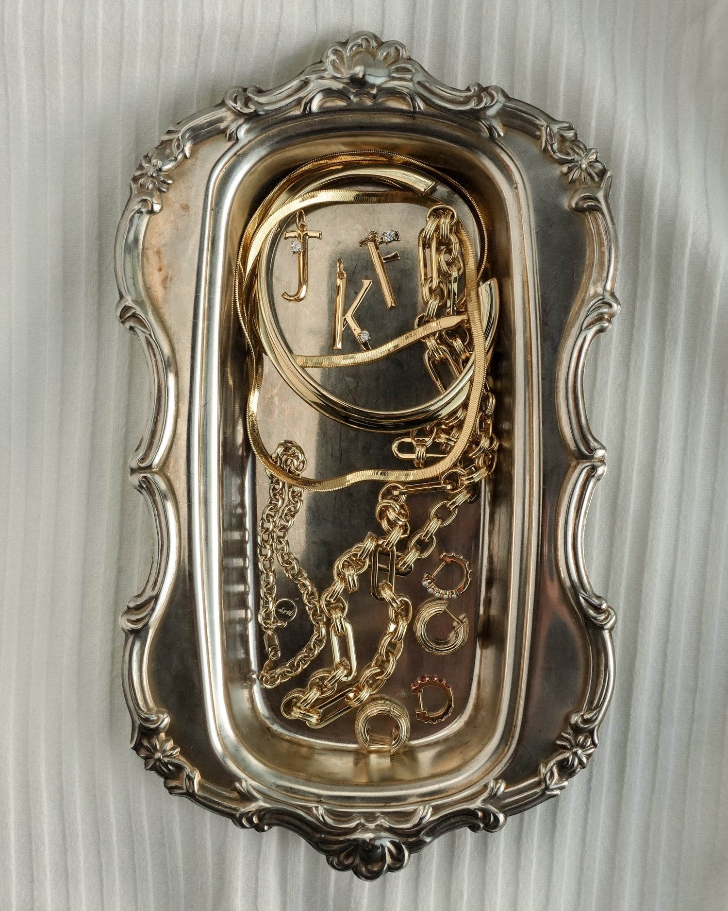 A decorative silver tray displays gold jewelry—including necklaces, bracelets, rings, and the Mod + Jo Eloise Letter Charm—arranged on a light pleated fabric background.