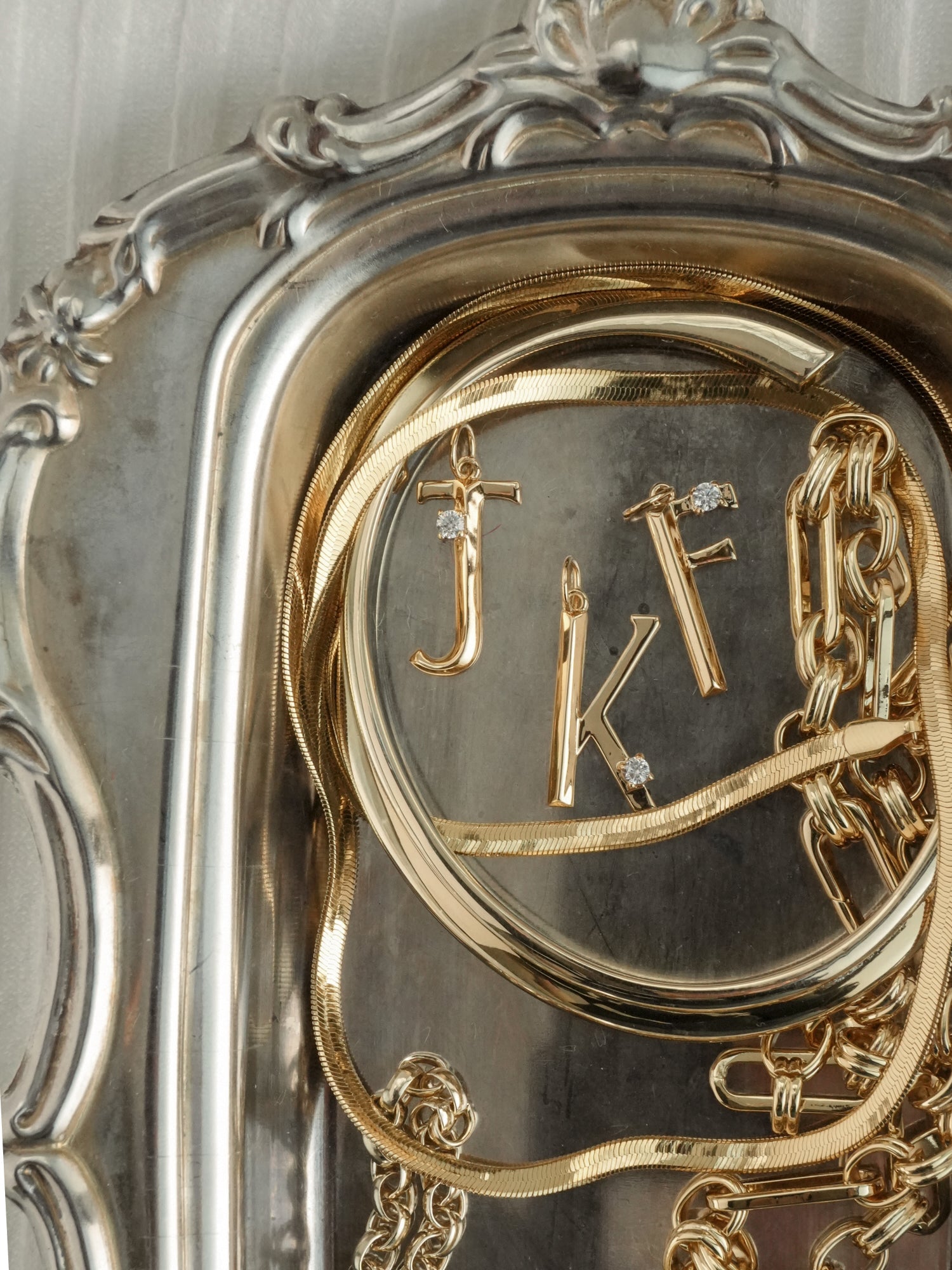 Mod + Jo's Eloise Letter Charms—J, K, and F—in gold with some featuring sparkling cubic zirconia, are displayed on a decorative silver tray.