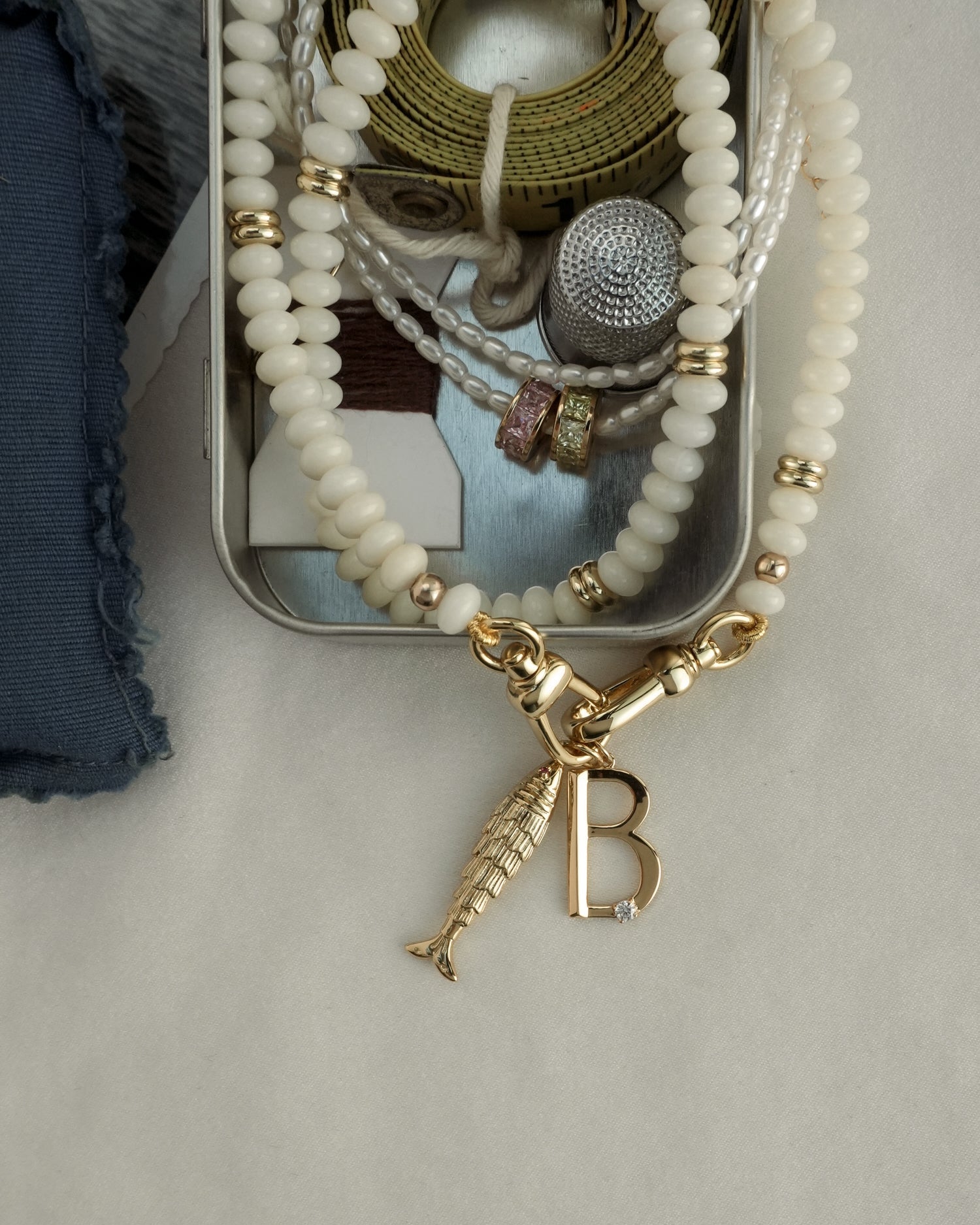 The Merit Gemstone Necklace by Mod + Jo is displayed on a beige surface beside a tin containing beaded bracelets, a watch strap, mother of pearl accents, and other 14k gold-filled jewelry.