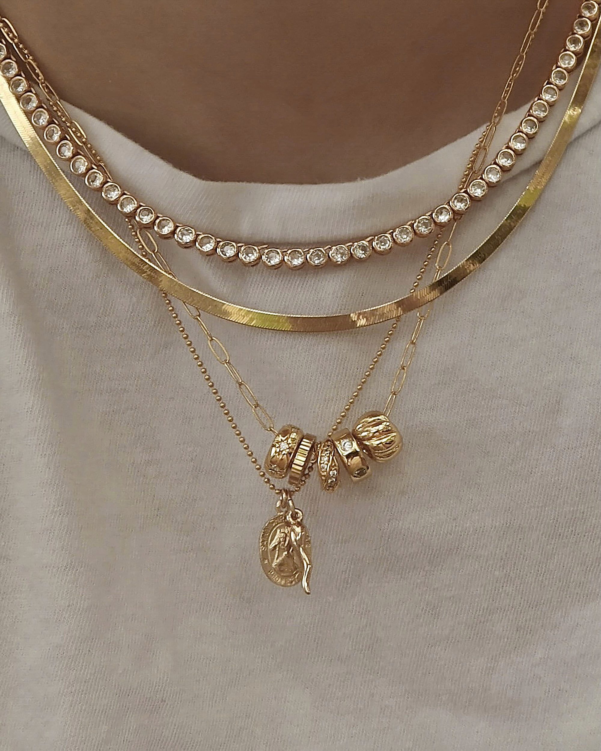 A person wearing layered gold necklaces, including a chain with clear gemstones, a smooth curved chain, and two thinner chains—one with a coin pendant and another with four gold charms, over a white shirt.