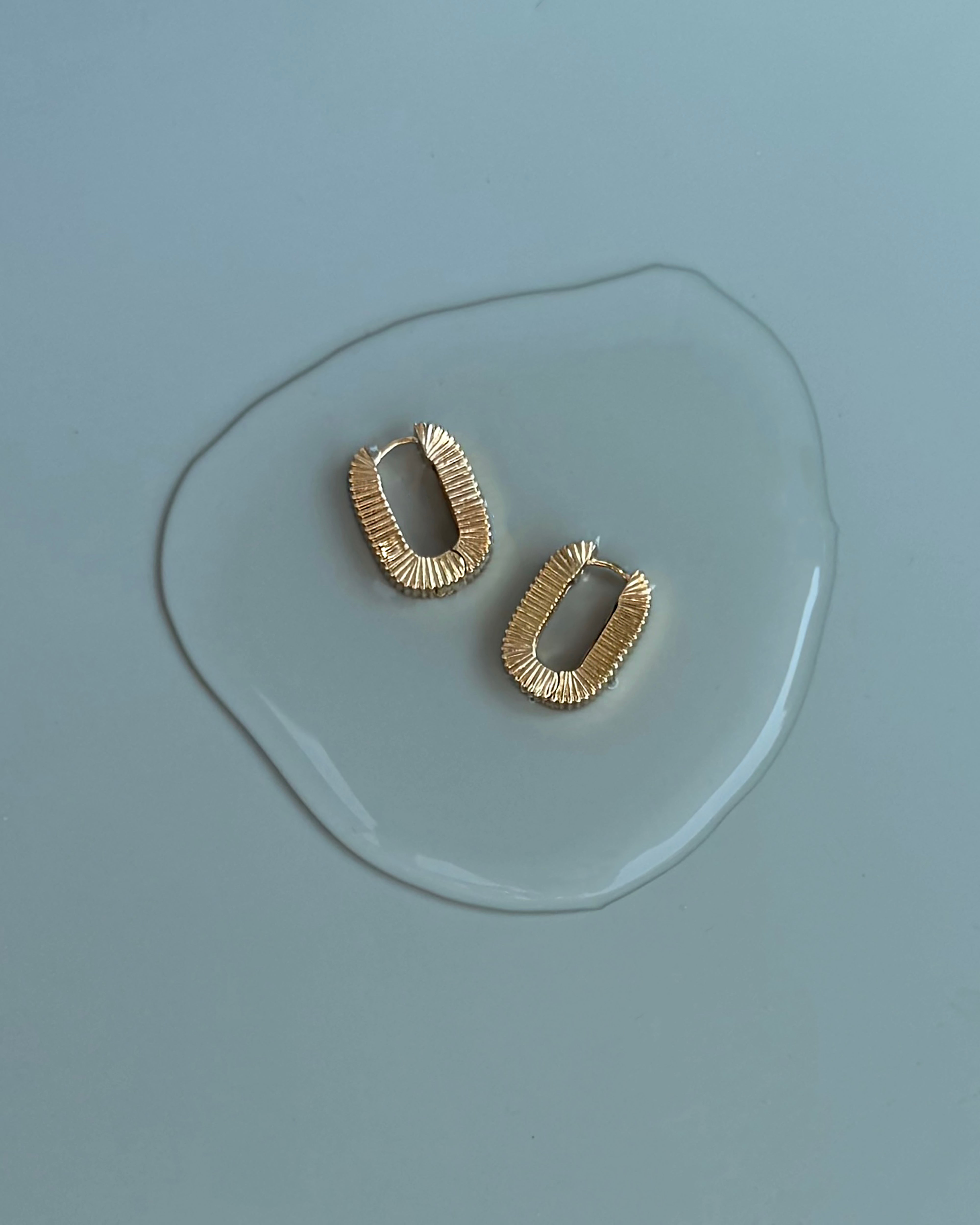 Two Elly Fluted Hoops by Mod + Jo, featuring rectangular shapes and textured gold surfaces, rest on a light blue surface, partially encircled by a clear puddle of water.