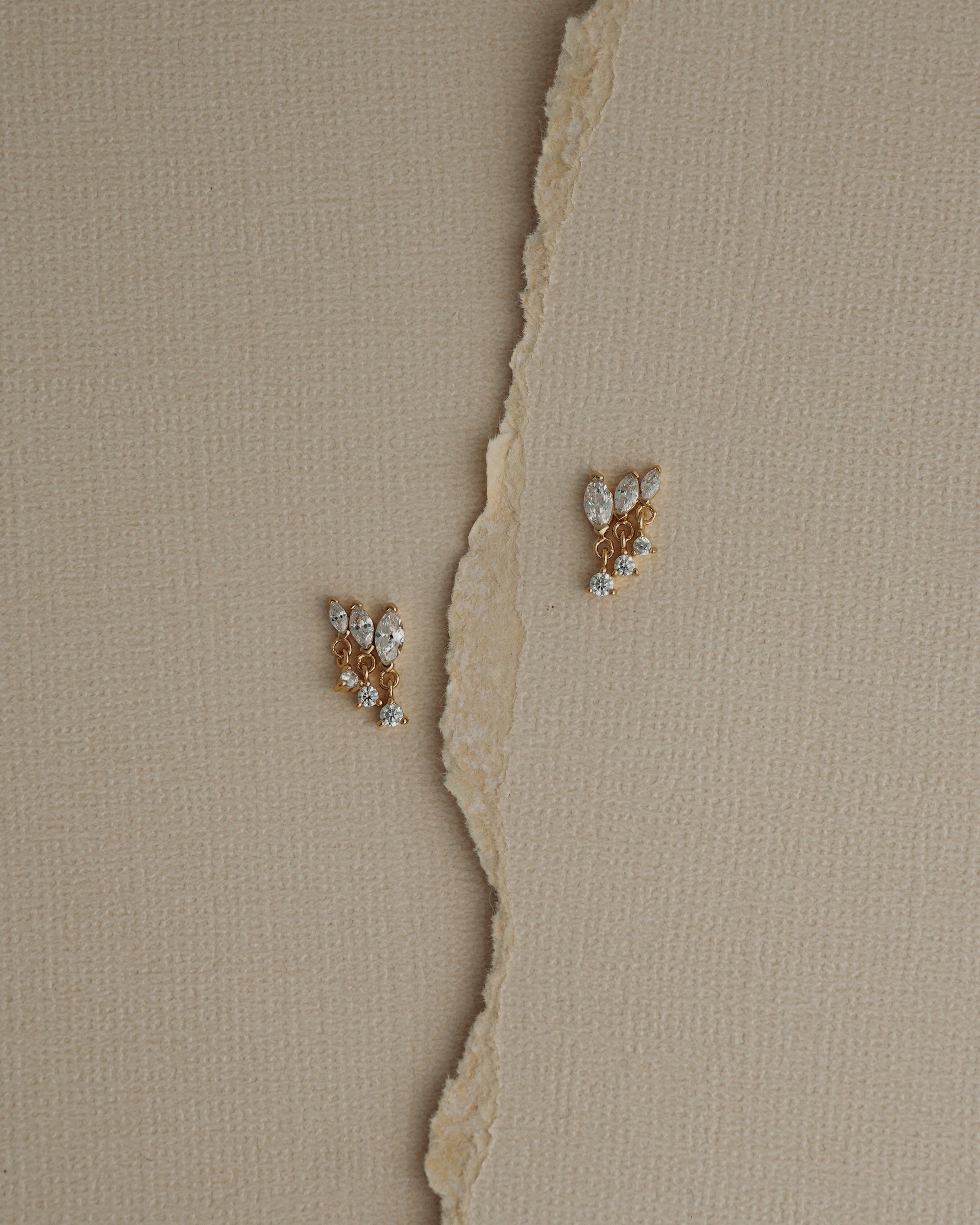 Two Gabby Marquise Dangle Studs by Mod + Jo, gold plated and adorned with sparkling cubic zirconia, rest on a textured beige surface, separated by a torn vertical strip of paper.