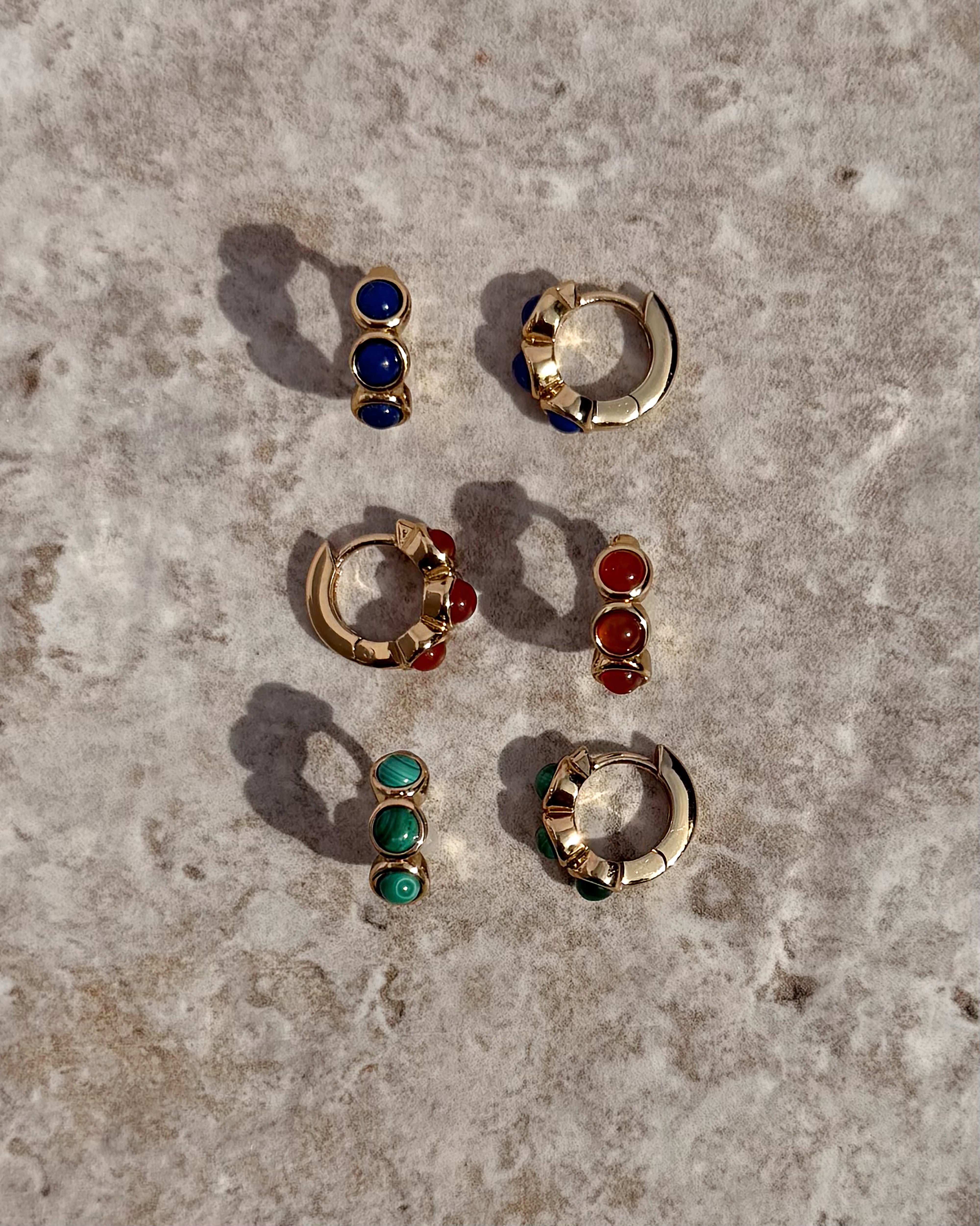 Three pairs of Mod + Jo’s *Preorder* Corynn Gemstone Huggies are shown on stone, each 18k gold plated and adorned with two gems—one pair with blue stones, one with genuine red agate, and another with green stones for a vibrant trio.