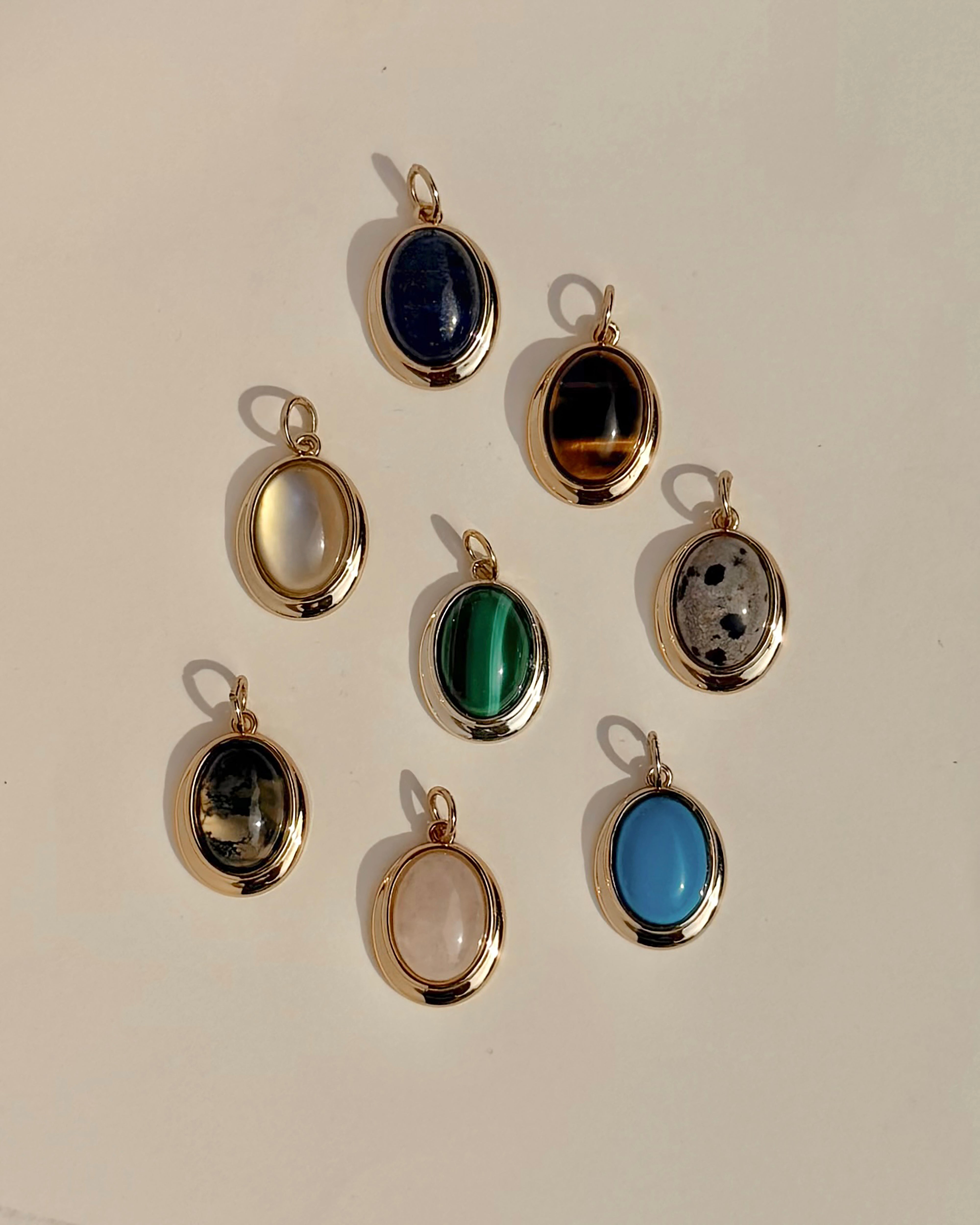 Eight exquisite Rory Gemstone Charms by Mod + Jo, each framed in 14k gold, are elegantly arranged on a light surface. Every charm showcases a distinct gemstone hue—serene blue, vibrant green, mysterious black, luminous white, gentle pink Rose Quartz symbolizing self-love, and uniquely patterned multicolored stones.
