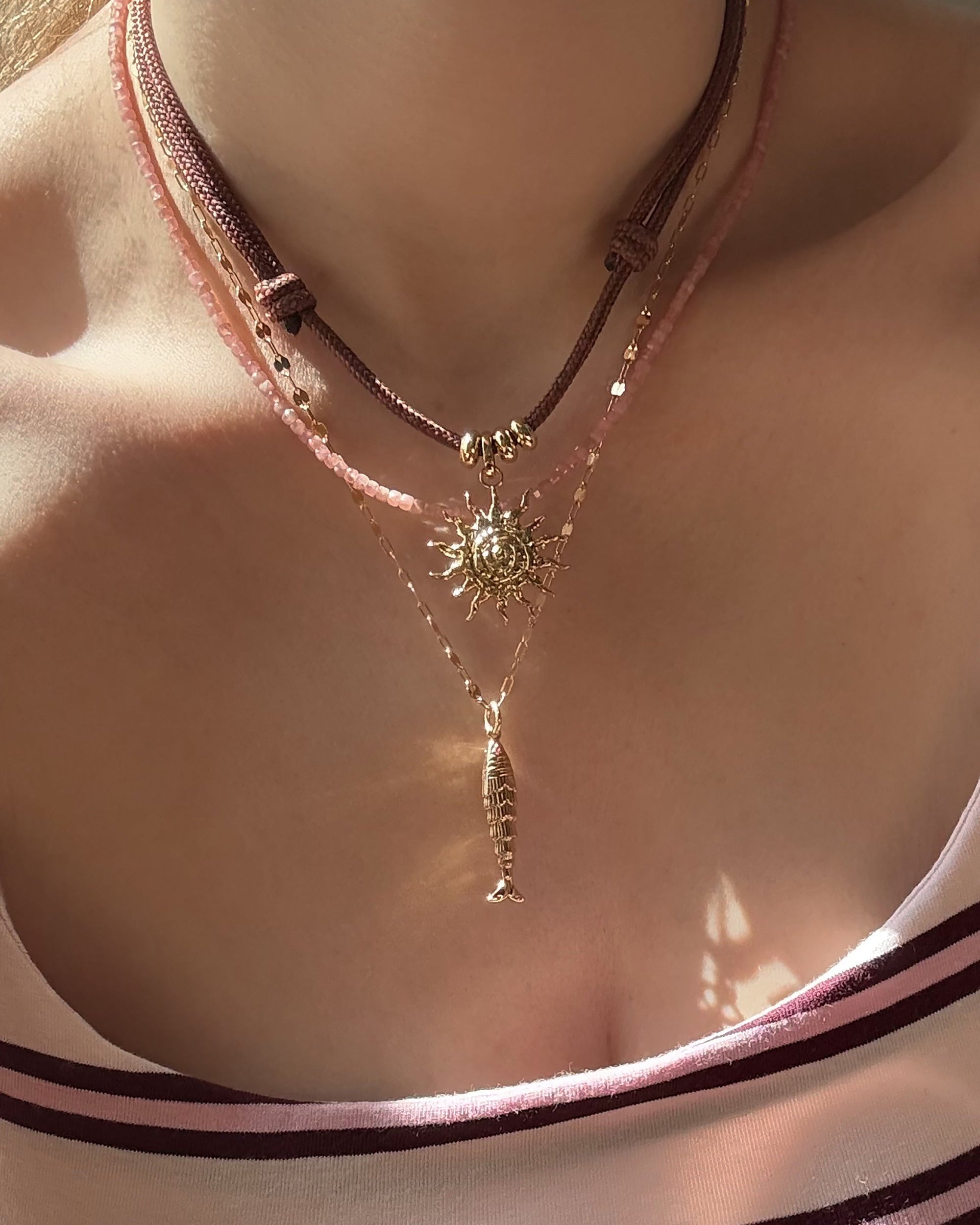 A close-up of a person’s neck wearing three layered necklaces: Mod + Jo’s Gillian Spiral Sun Charm on a brown cord with gold beads, a pink beaded choker, and an 18k gold-plated fish pendant chain. The person wears a striped top.