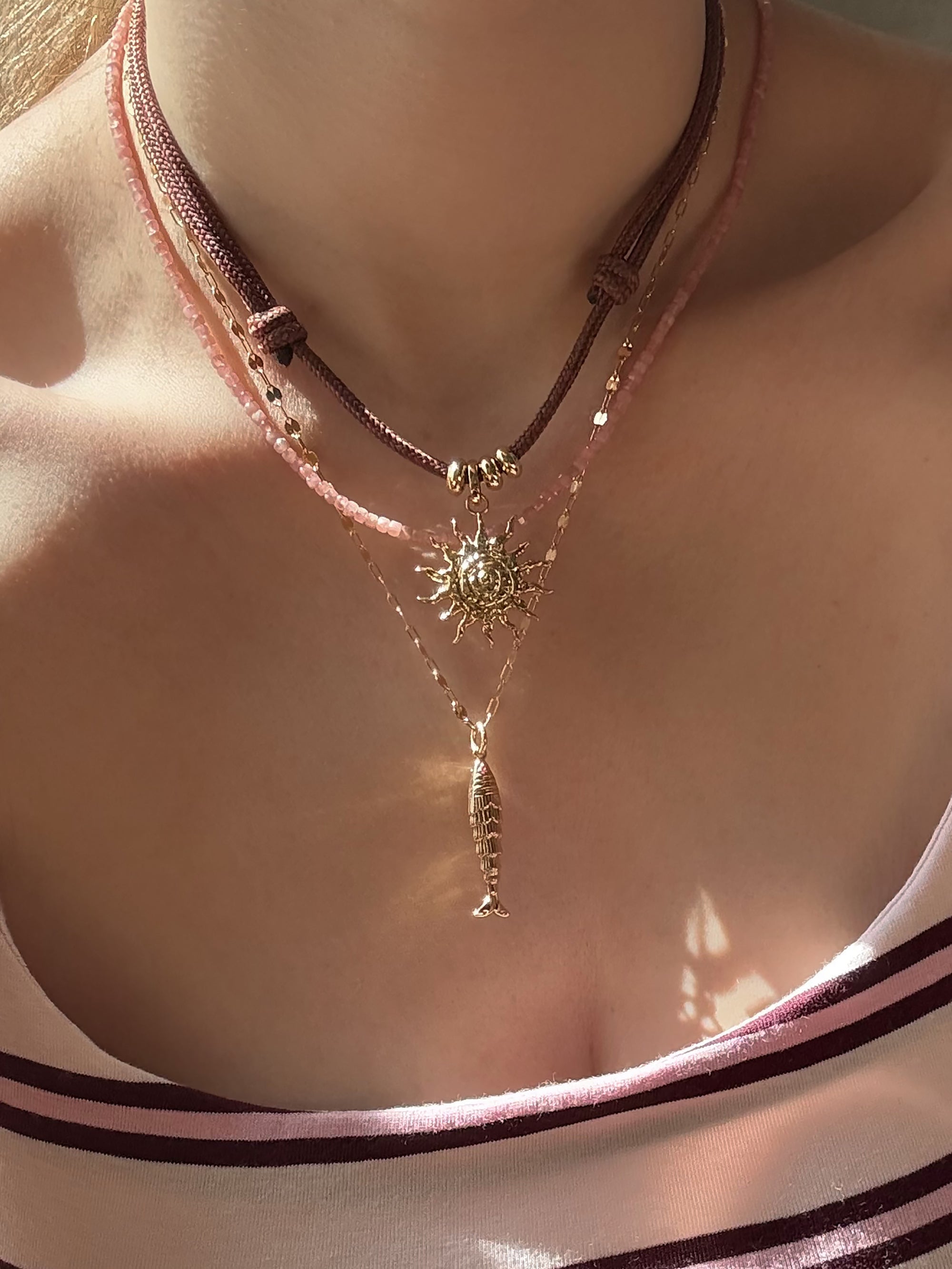 A close-up shows a person in a pink and maroon striped top wearing the Mod + Jo Tommy Cord Necklace layered with two other necklaces: a gold sun pendant, a fish pendant, and a pink beaded choker.