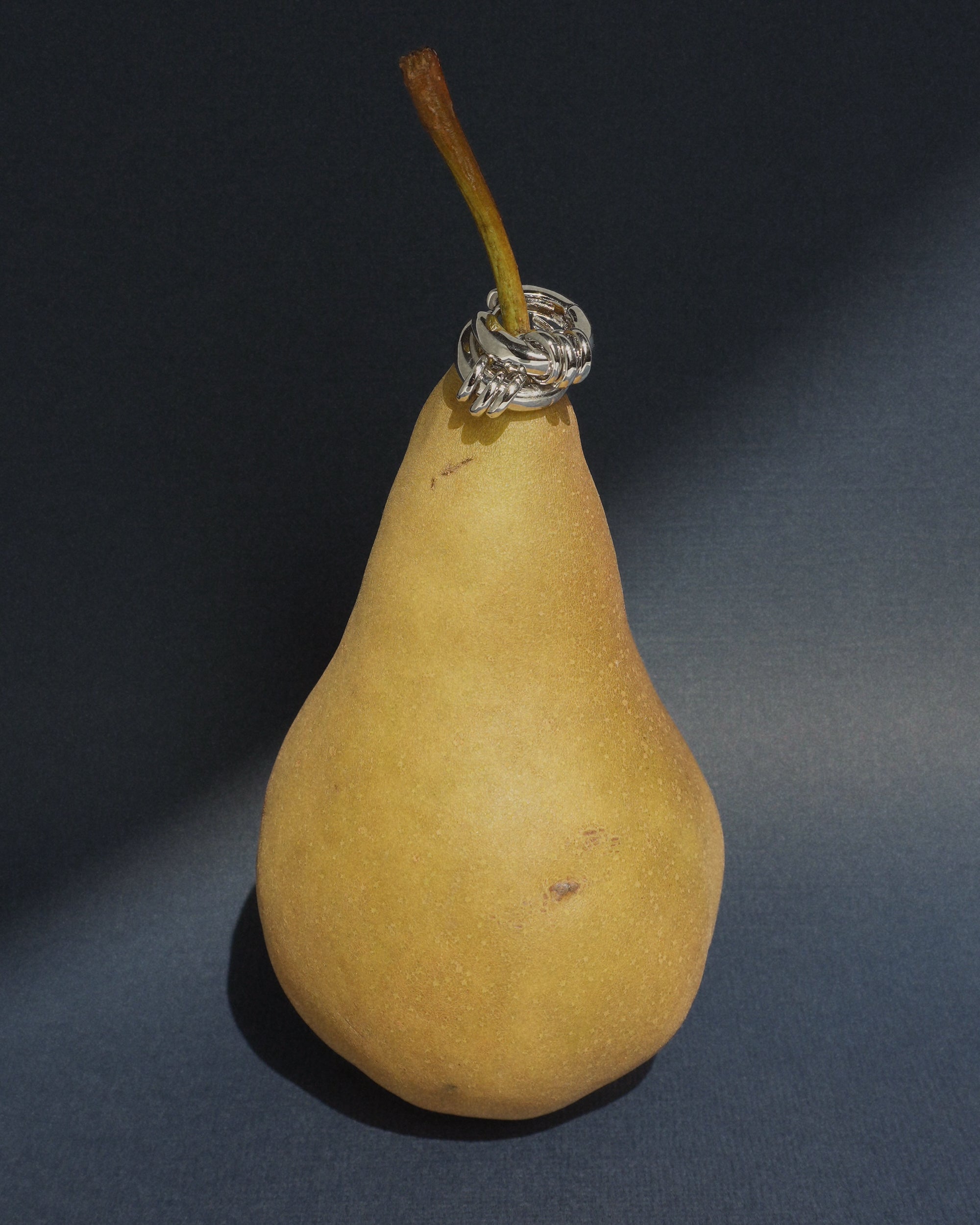 A yellow pear stands upright on a dark surface, its stem adorned with a stack of silver rings reminiscent of statement huggie earrings, evoking the chic design of the rhodium-plated Grande Levi Huggies by Mod + Jo.