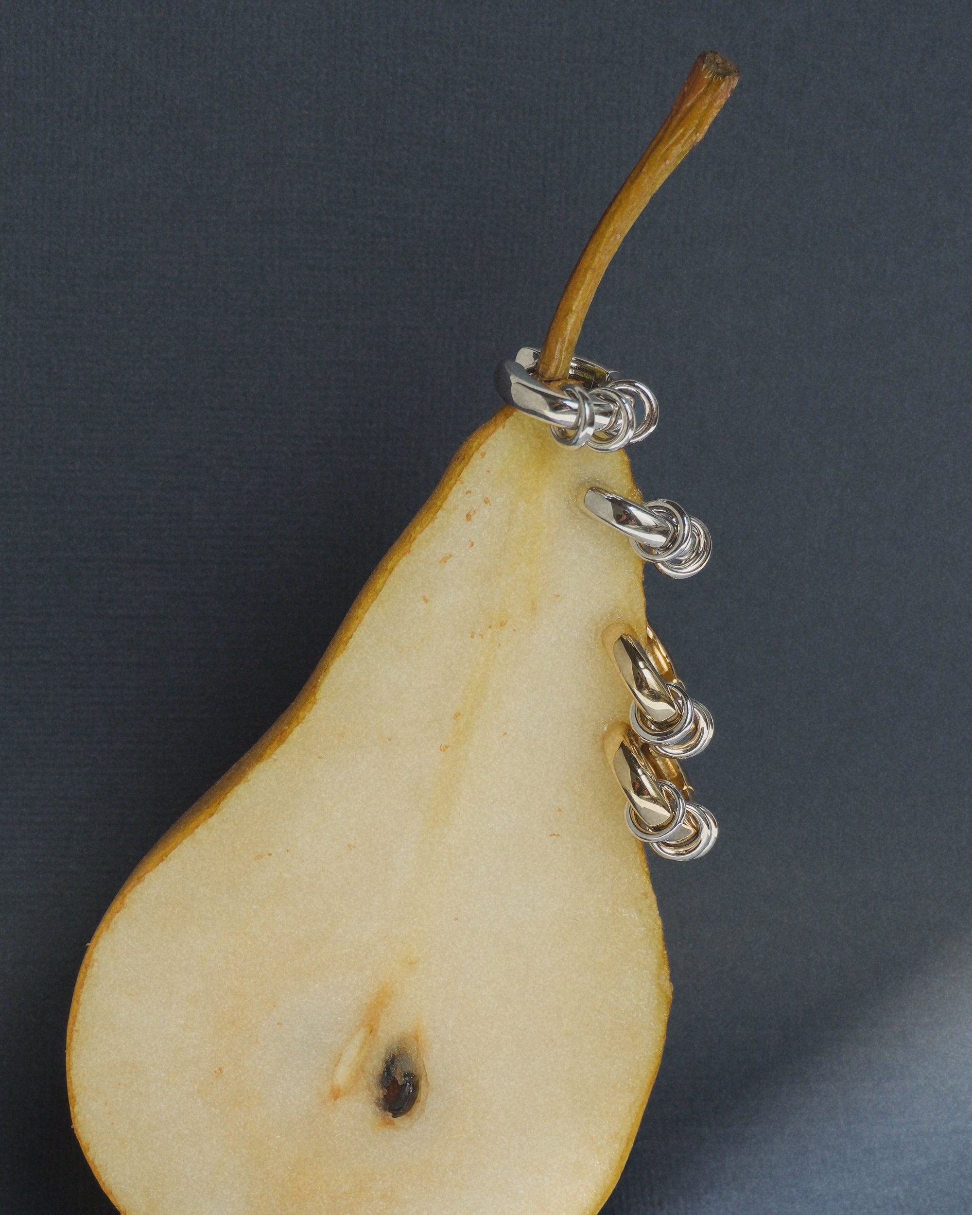 A sliced pear half with its stem attached, embellished with small metal chain rings along one side—evocative of the graceful links featured in Mod + Jo’s Grande Levi Huggies, gold-plated huggie earrings that are carefully crafted to be nickel and lead free—rests on a dark textured background.