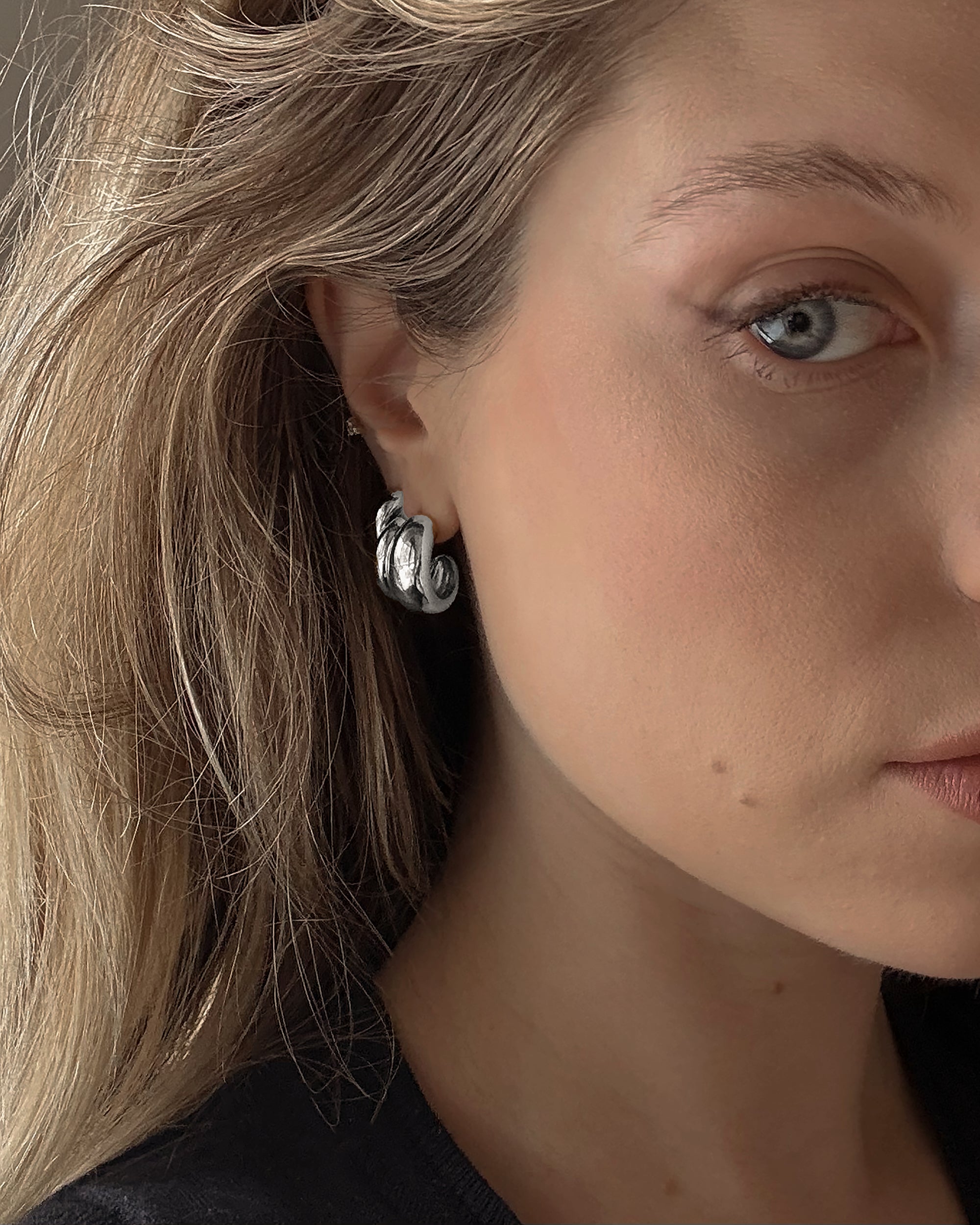 Close-up of a woman with light skin and blonde hair wearing Mod + Jo's Grande Sofie Volume Hoops. Only half her face and one blue eye are visible, softly lit to highlight the chunky rhodium plated silver earrings.