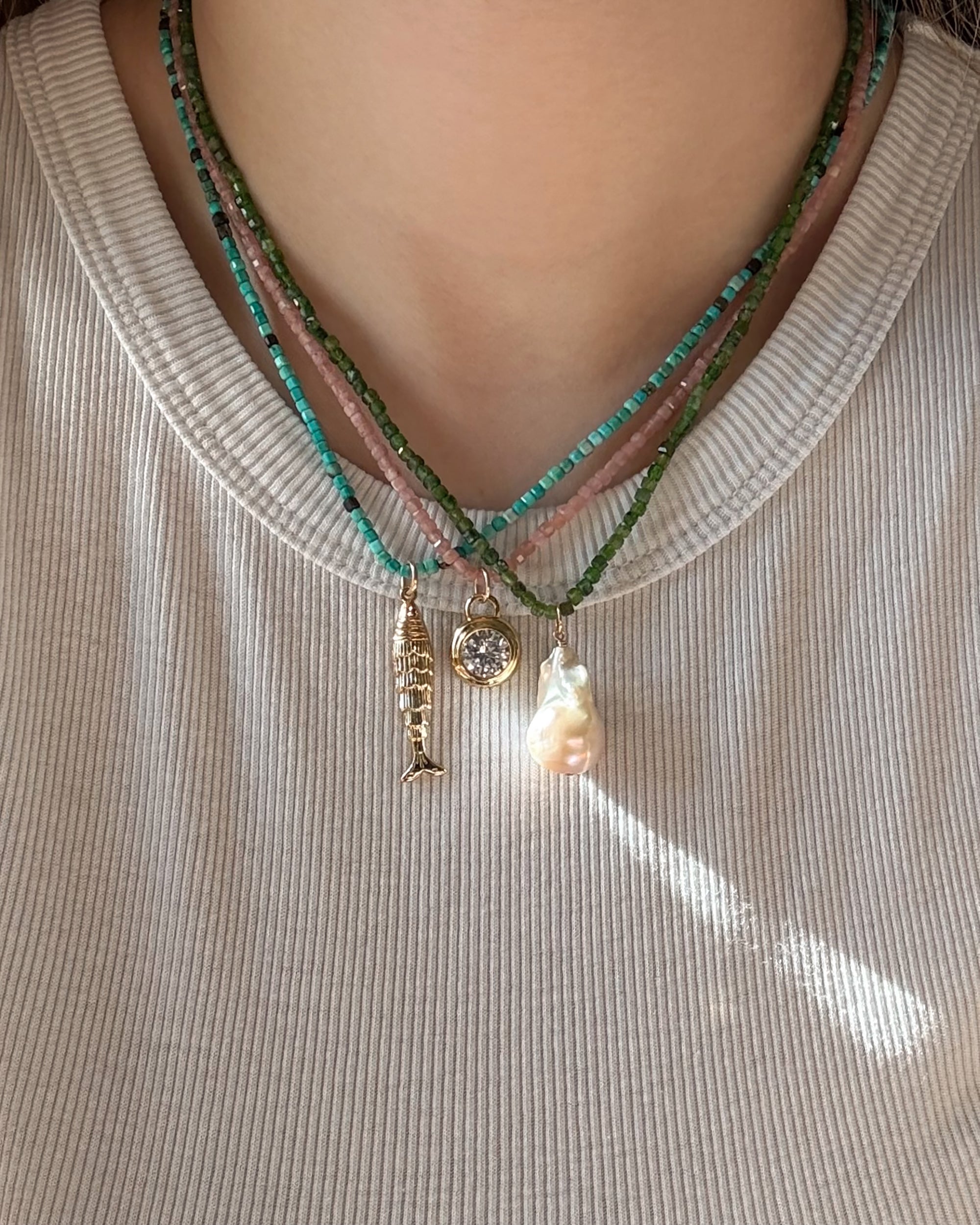 A close-up of a person in a light ribbed shirt wearing three Mod + Jo Merit Gemstone Necklaces, each featuring unique pendants—a diver, a round gem, and an irregular pearl—adorned with green and turquoise beads.