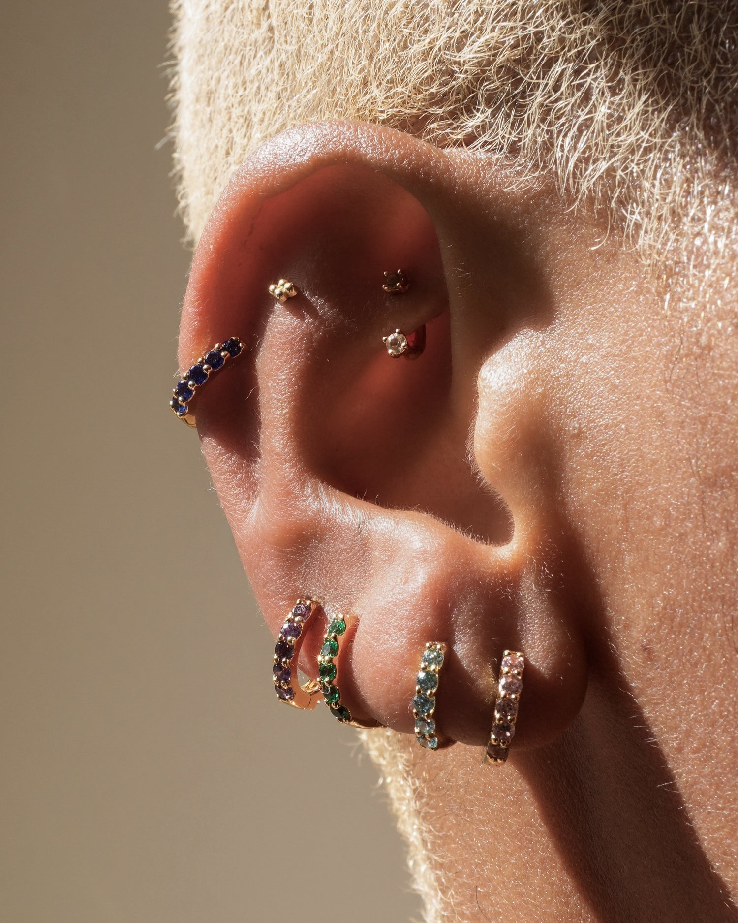 A close-up of a person with short, textured, light hair wearing Mod + Jo Harlow Reversible Birthstone Huggies in 18k gold plating, featuring blue, green, and light pink gemstones.