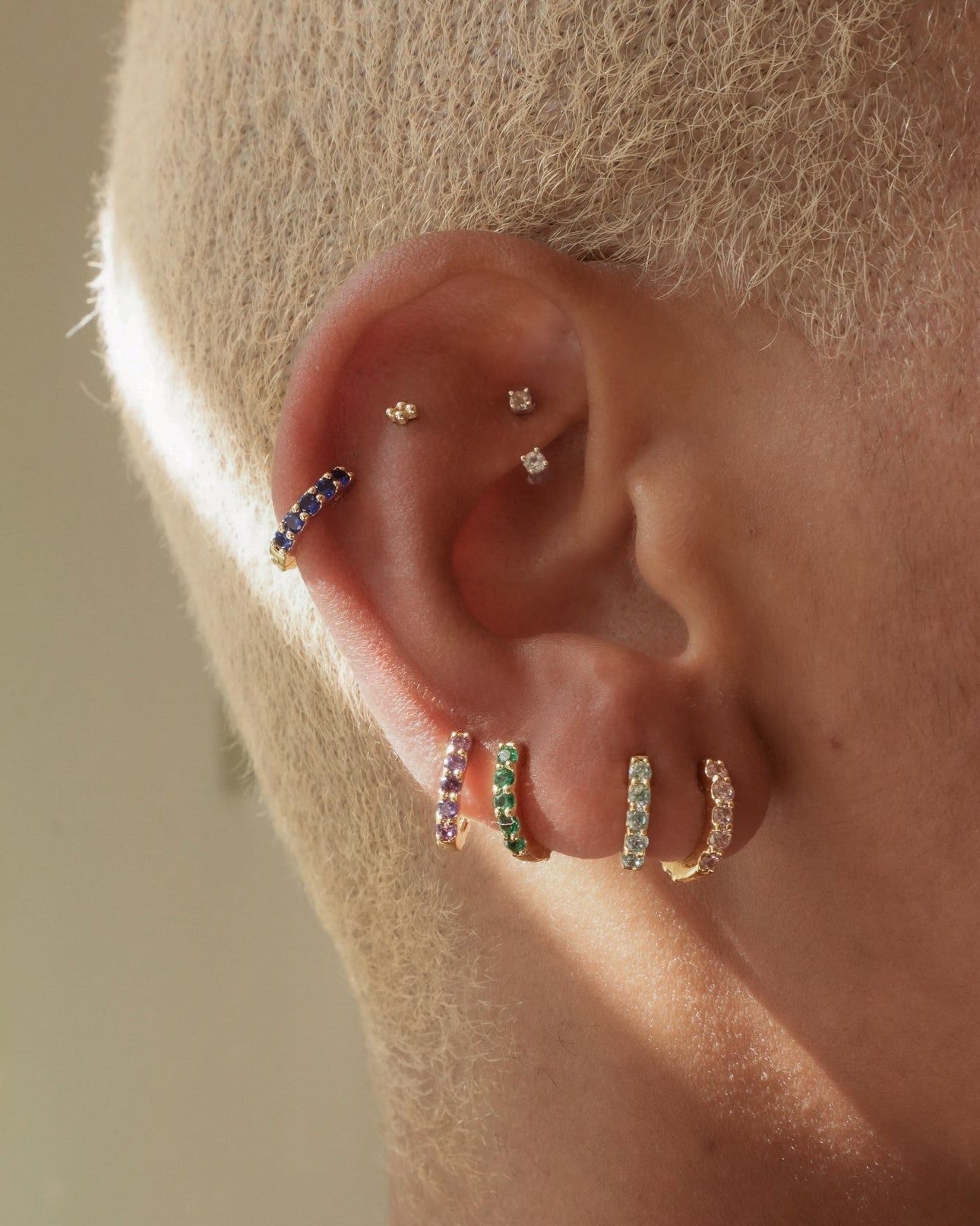 A close-up of an ear with multiple piercings, featuring Mod + Jo Harlow Reversible Birthstone Huggies in various colors and two small diamond studs. The person has short, bleached blonde hair.
