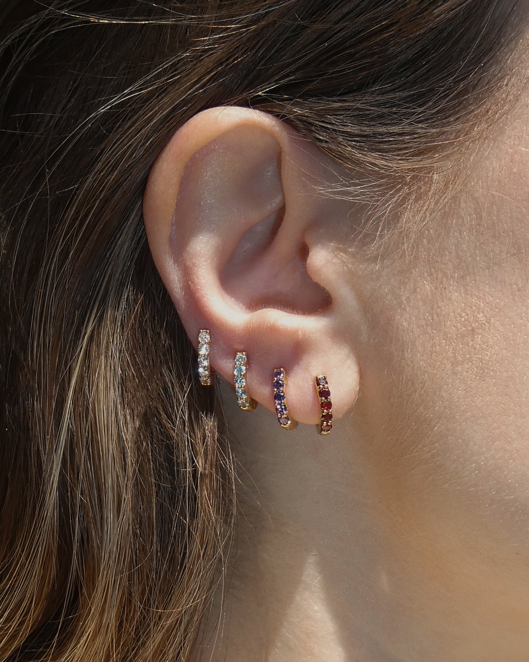 Close-up of a woman's ear wearing four Mod + Jo Harlow Reversible Birthstone Huggies with white, green, blue, and reddish-purple gems, arranged in a graduated line along the ear.