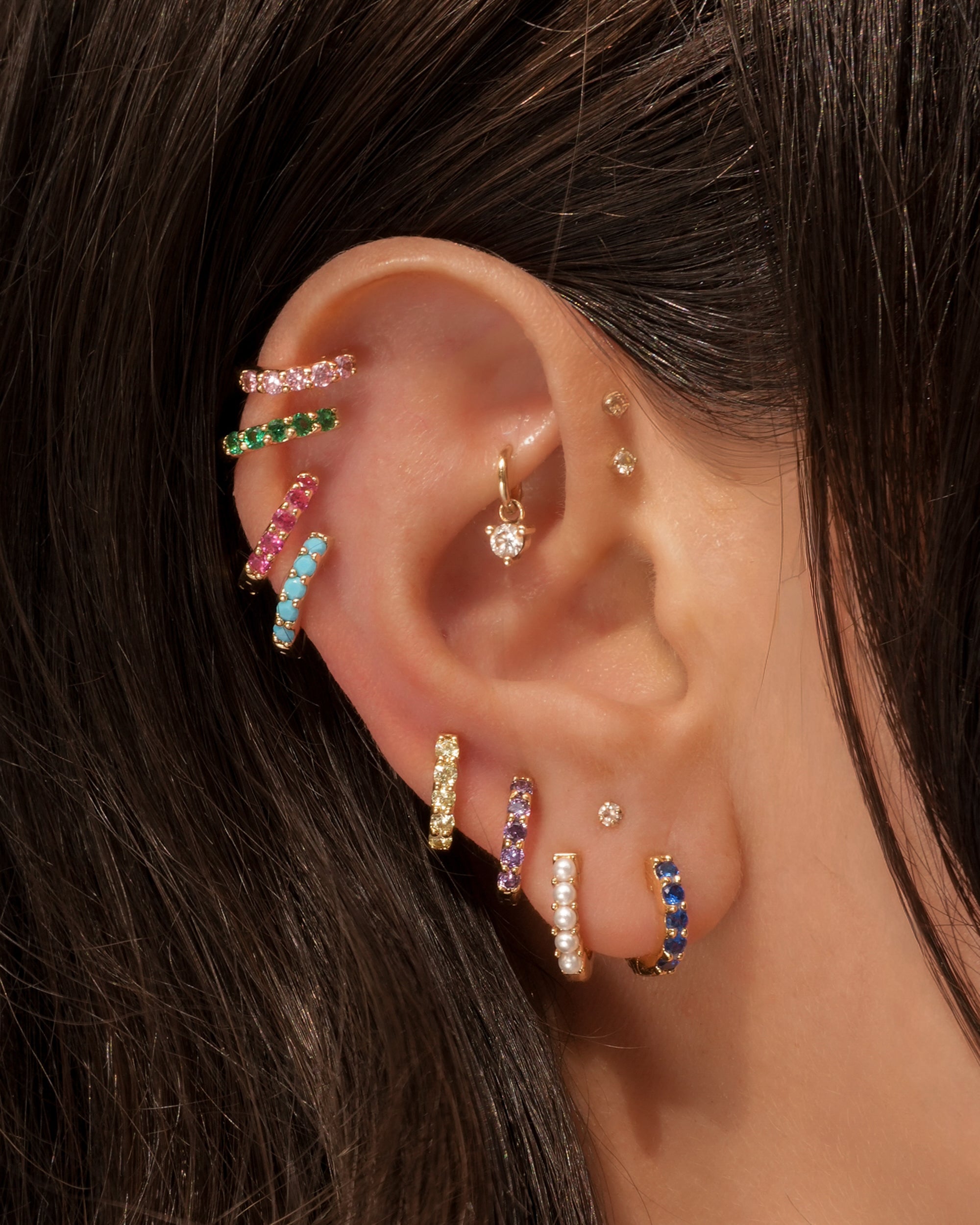 A close-up of a person’s ear wearing Mod + Jo’s Harlow Reversible Birthstone Huggies and small 18k gold plated studs, with the jewelry shown against dark brown hair.