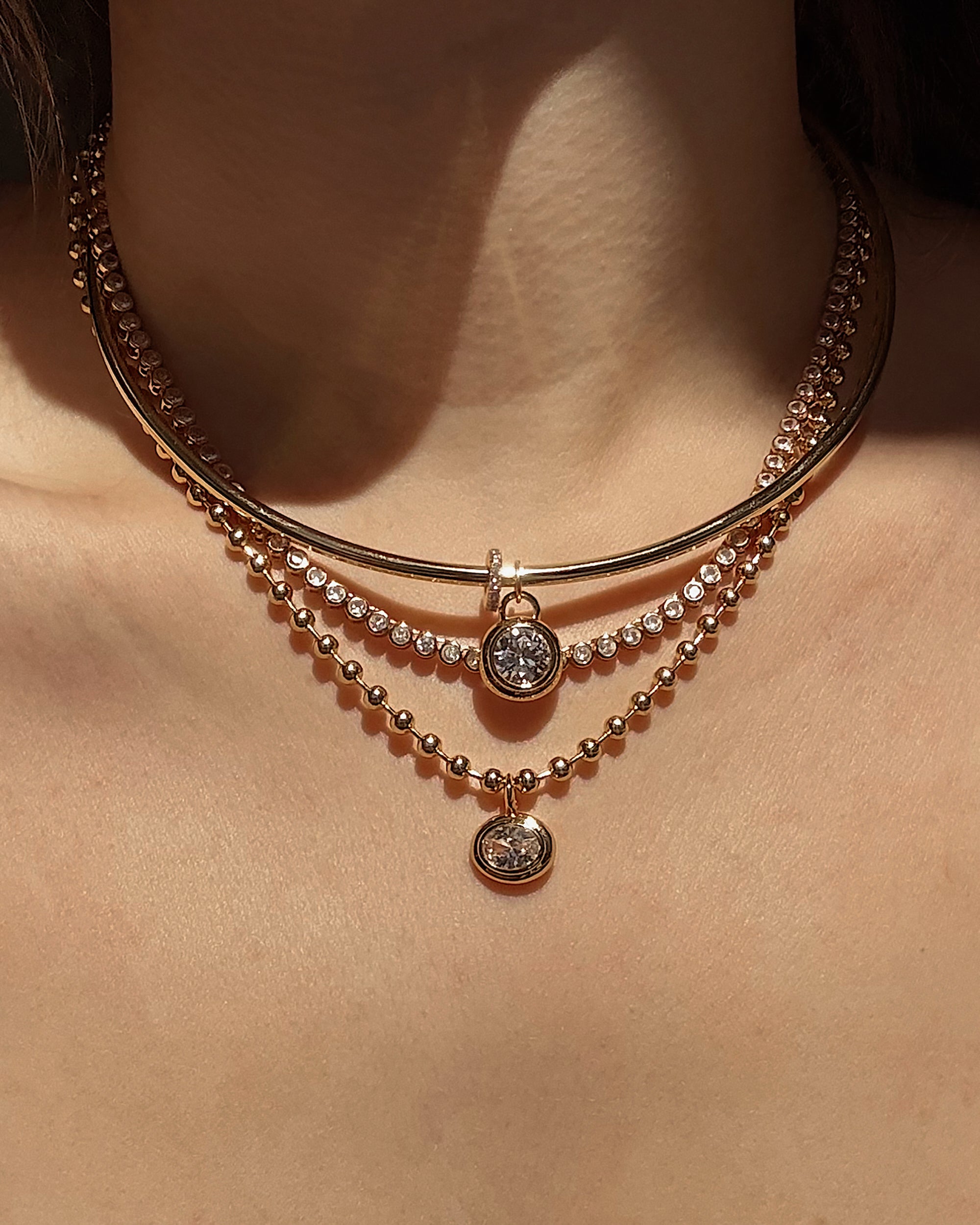 A close-up of a woman's neck wearing three layered gold necklaces by Mod + Jo: a solid choker, a crystal chain, and two pendant necklaces with round gemstones and the Tove Sapphire Bezel Charm featuring a sparkling lab-grown sapphire.