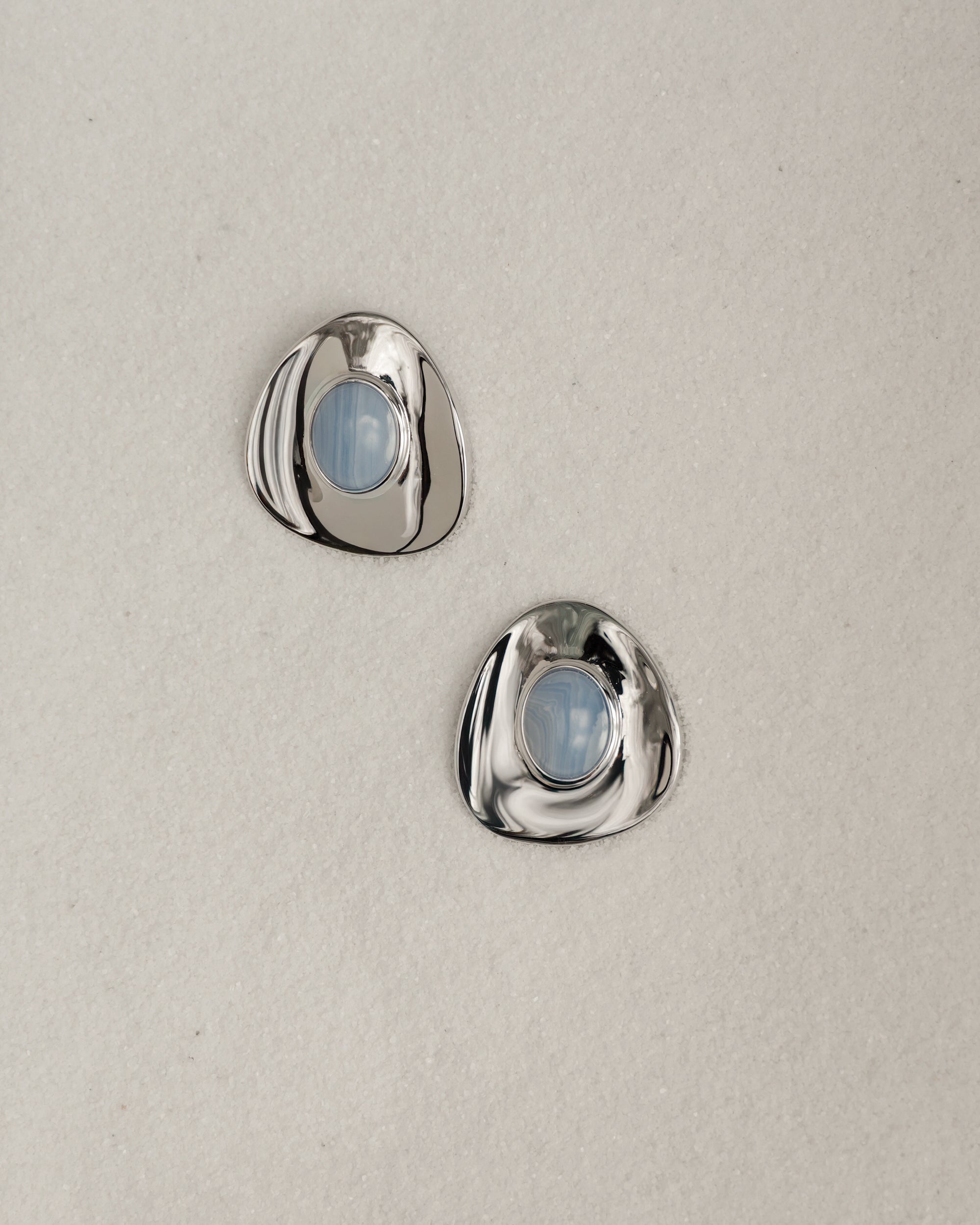 The Mod + Jo Hunter Statement Studs are two shiny, irregular silver earrings with round blue chalcedony centers, displayed on a light beige textured surface.